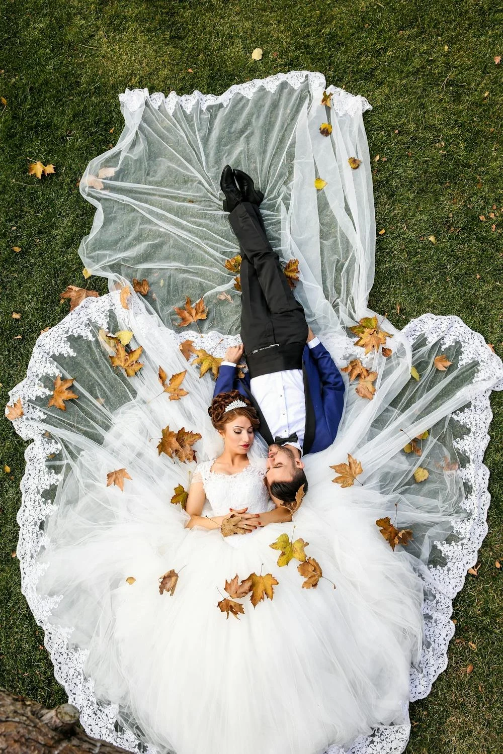 Bride in a white gown and groom in a blue suit lying on grass, surrounded by fallen autumn leaves, with lace fabric spread out around them.