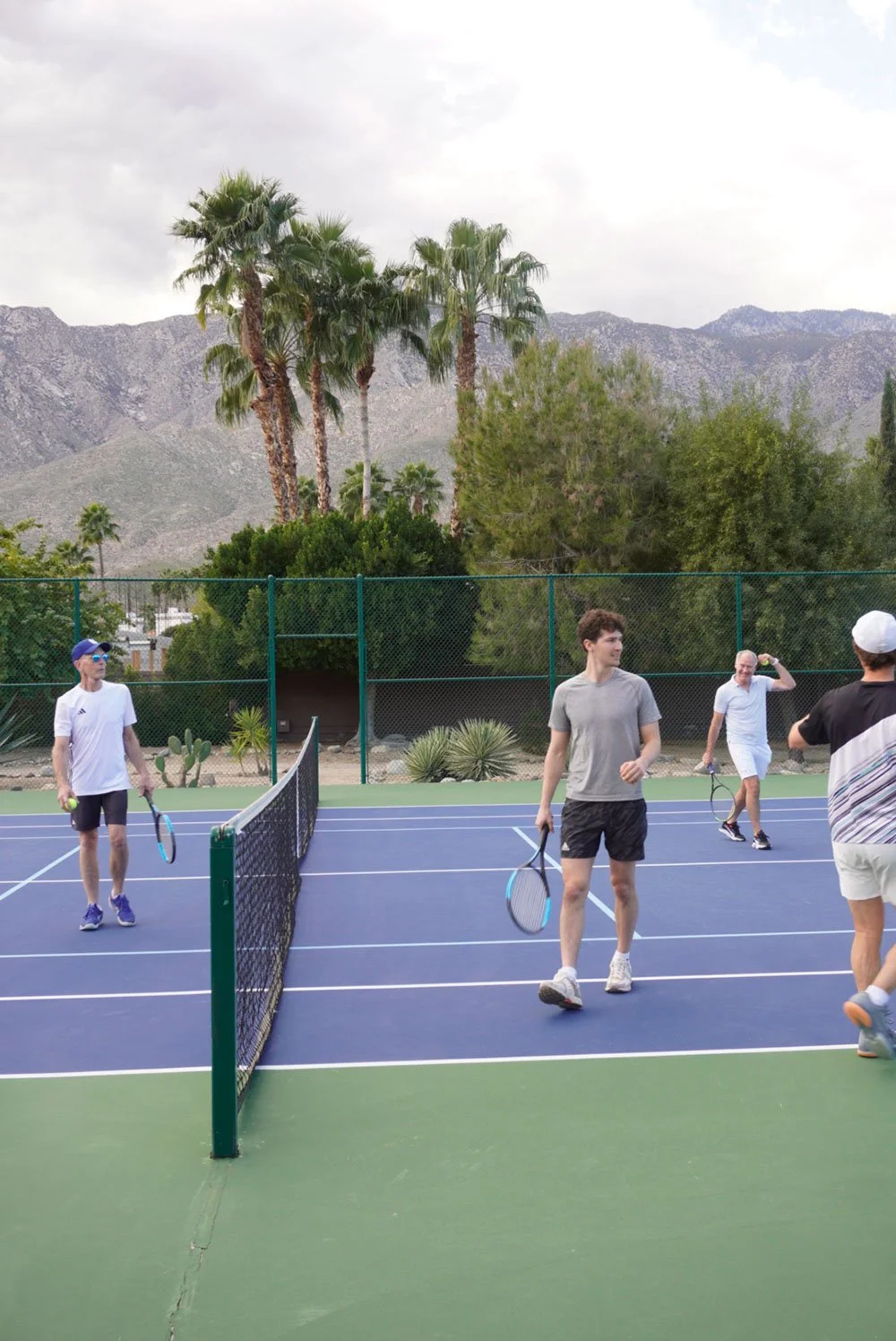 Group of men playing tennis on a court with palm trees and mountains in the background.
