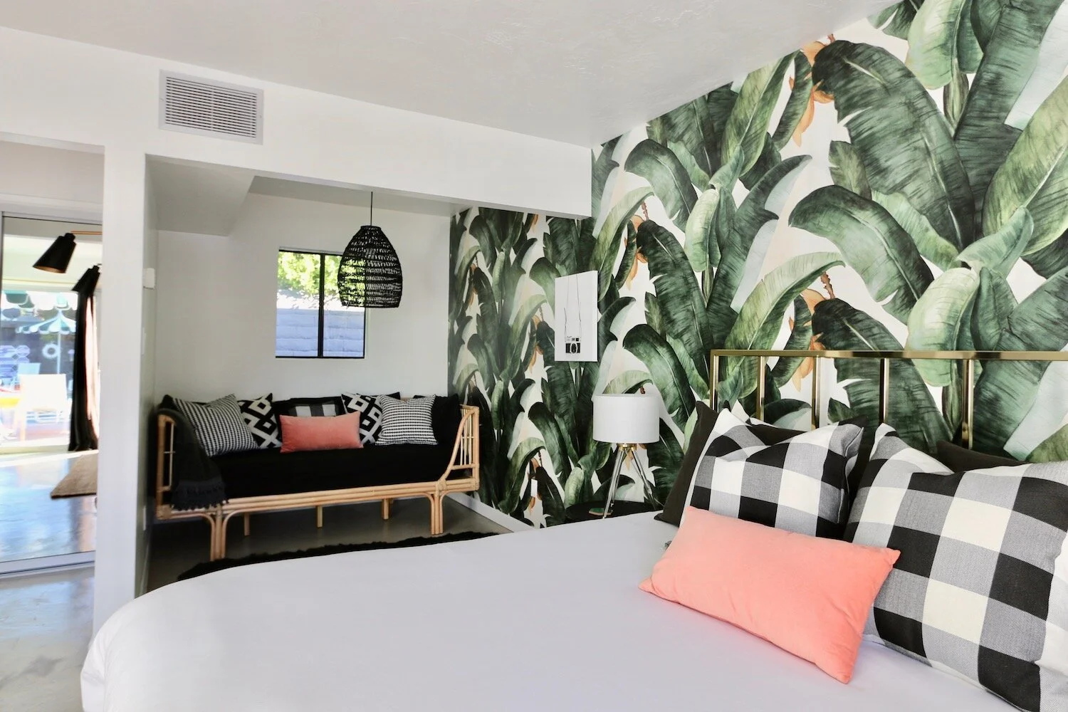 Modern bedroom with tropical leaf wallpaper, a bed with plaid and pink pillows, a rattan daybed with patterned cushions, and a black pendant light.