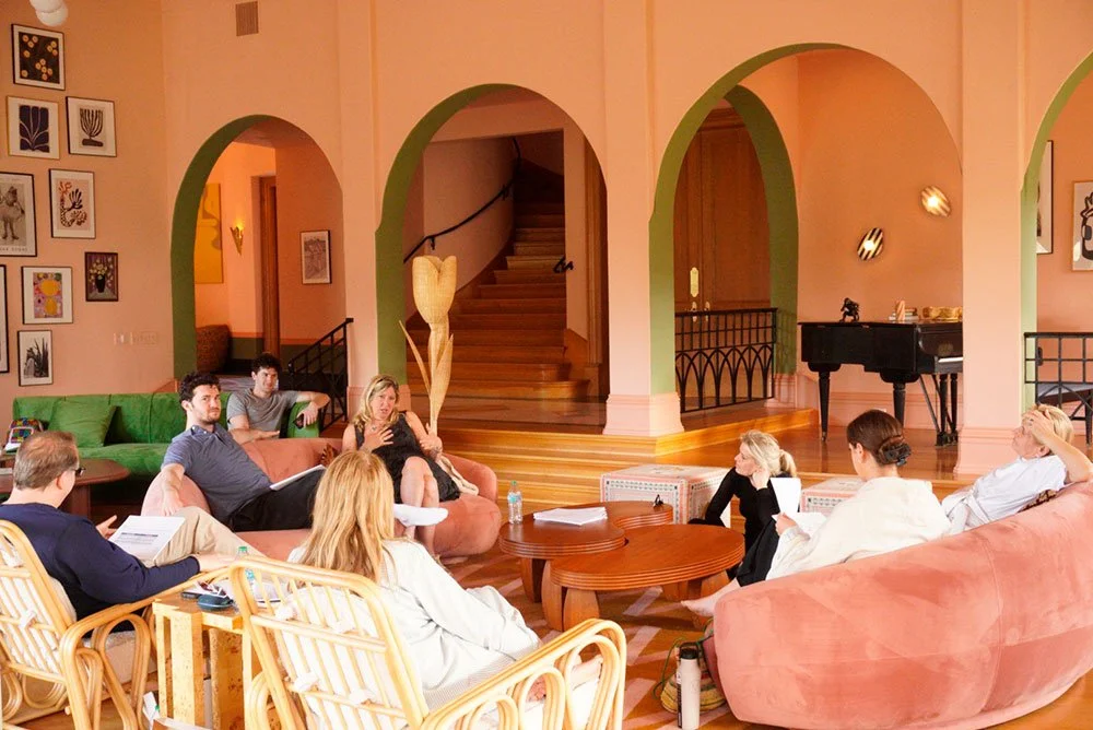 A group of people are gathered in a cozy, well-decorated living room for a meeting or discussion, with some sitting on sofas and chairs, and a grand piano in the background.