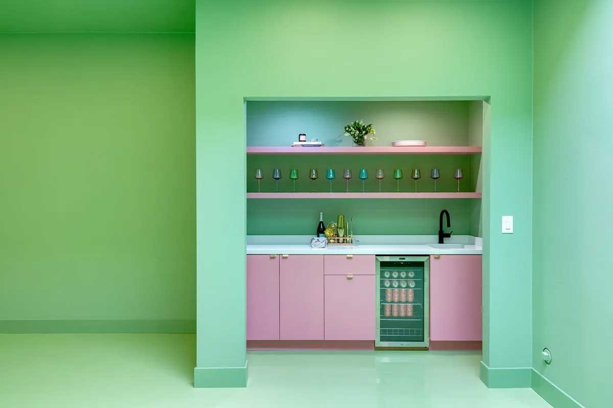 Modern kitchen alcove with pink cabinets, green walls, glass shelves holding colorful glasses, a small fridge, and a black faucet.