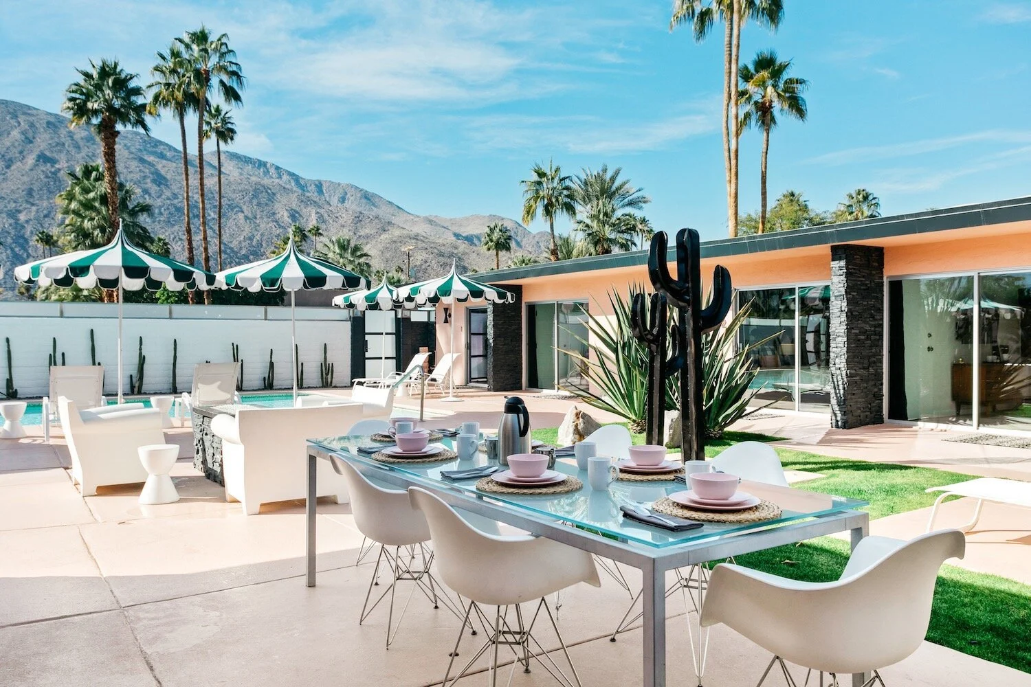 Outdoor patio with a glass dining table, white chairs, tableware, pool, umbrellas, plants, and mountain view.