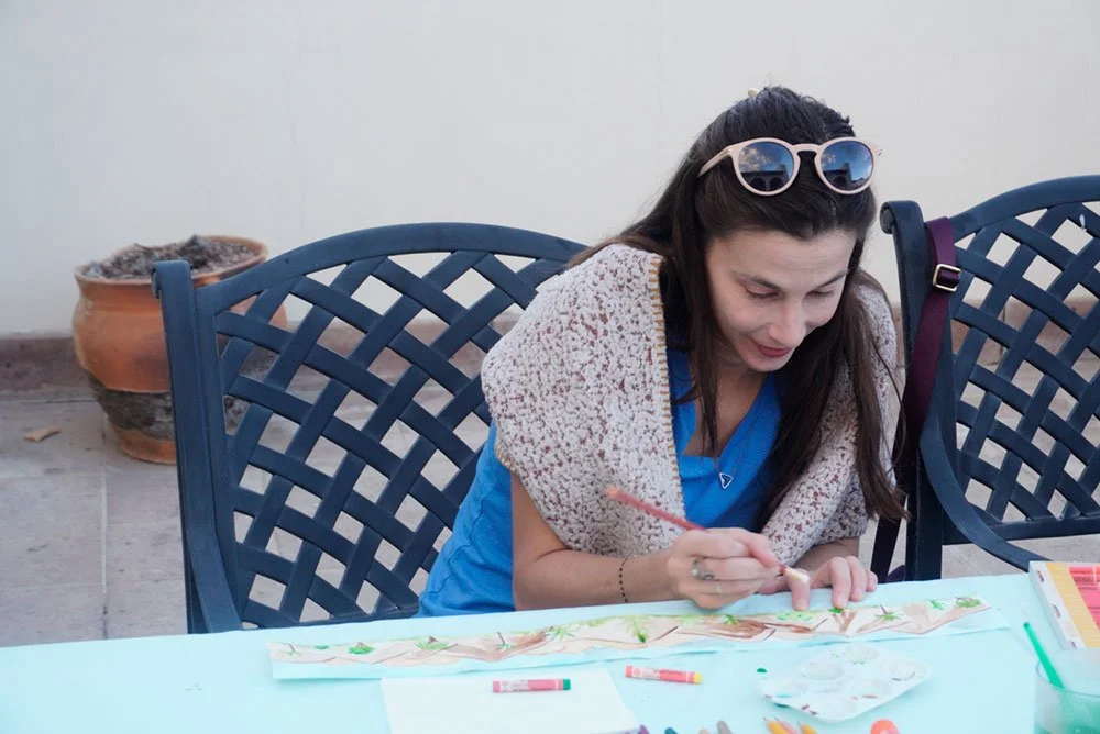 Woman sitting at a table outdoors, painting on paper with watercolors, wearing sunglasses on her head and a shawl.