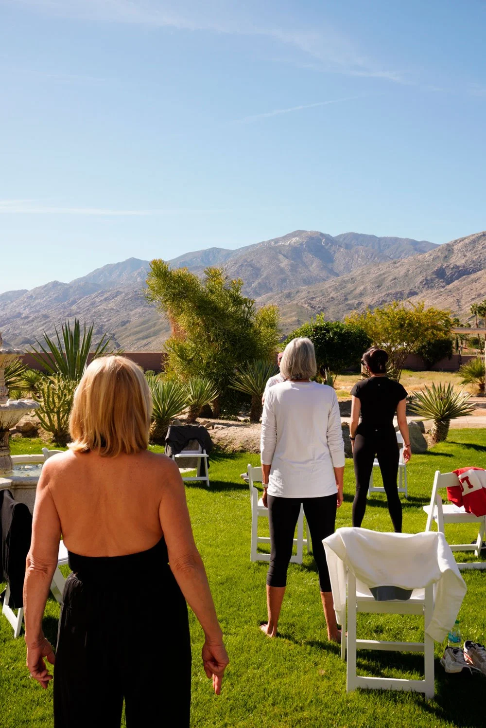 Women standing on a lush green lawn with mountains in the background, trees, and chairs, under a clear blue sky.