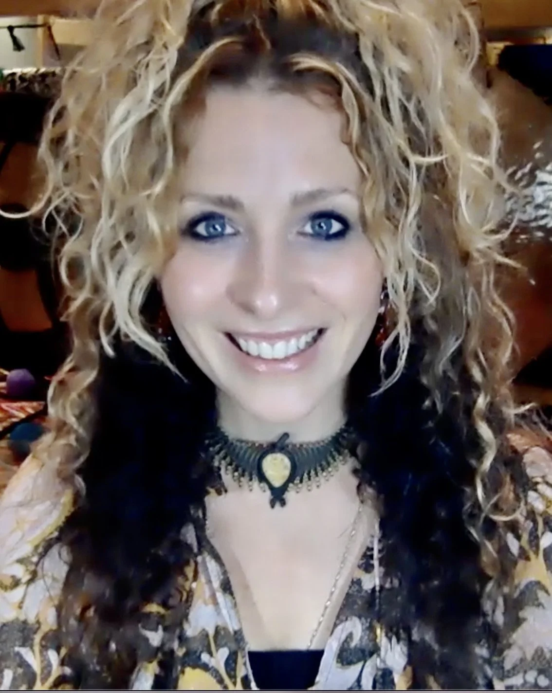Person with curly hair smiling, wearing a patterned top and a decorative necklace.