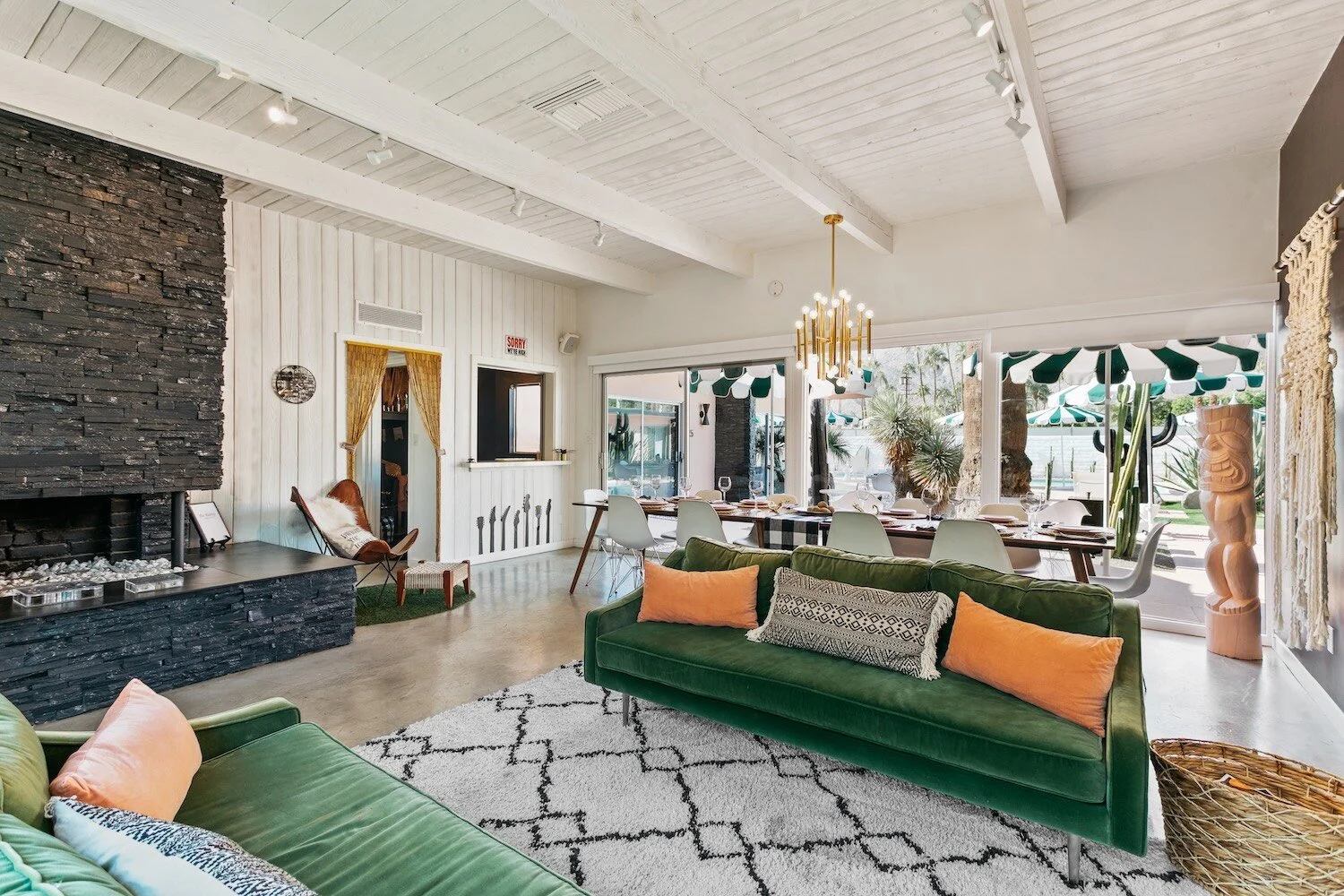 Modern living room with green sofas, a patterned rug, stone fireplace, large dining table, and glass doors opening to a patio with striped umbrellas.