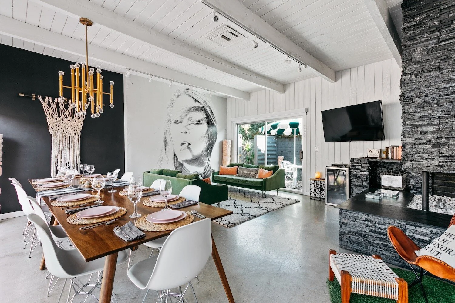 Modern living and dining area with a long wooden table set with plates and wine glasses, white chairs, a black stone fireplace, and a green couch. A large wall mural of a woman's face and a gold chandelier enhance the space. Sliding doors lead to an 
