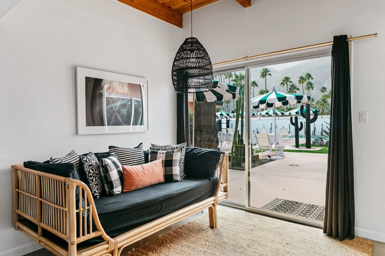 Interior of a modern living space with a wicker daybed, patterned pillows, and a black hanging light fixture. A large glass door opens to a patio with striped umbrellas, outdoor seating, and palm trees.