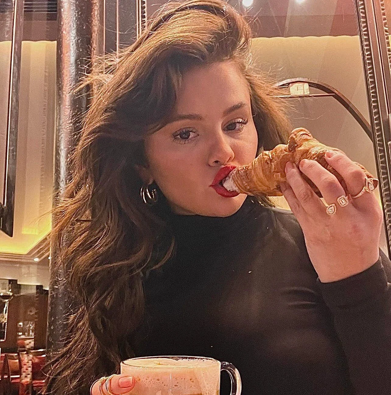 Mujer con cabello rizado oscuro, usando lápiz labial rojo, sosteniendo una pieza de pan o croissant, en un ambiente de restaurante o cafetería, con una taza con bebida en primer plano.
