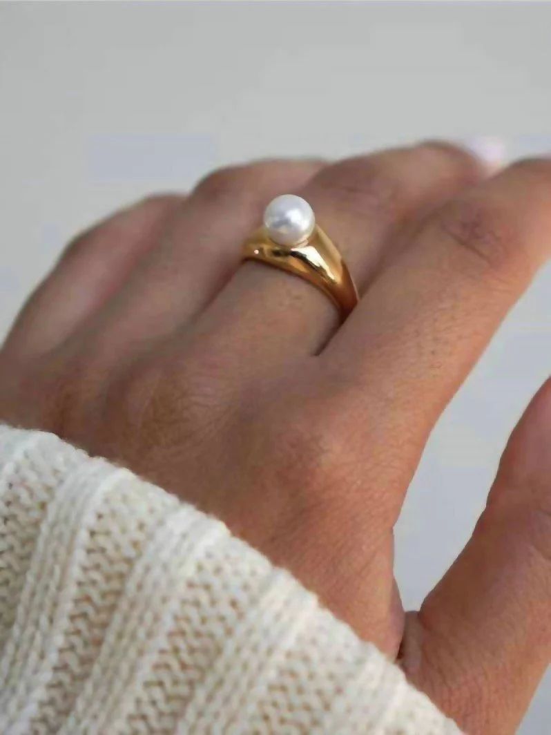 Anillo de oro con perla en el dedo de una mano con piel de tono medio y ropa de punto en color crema.