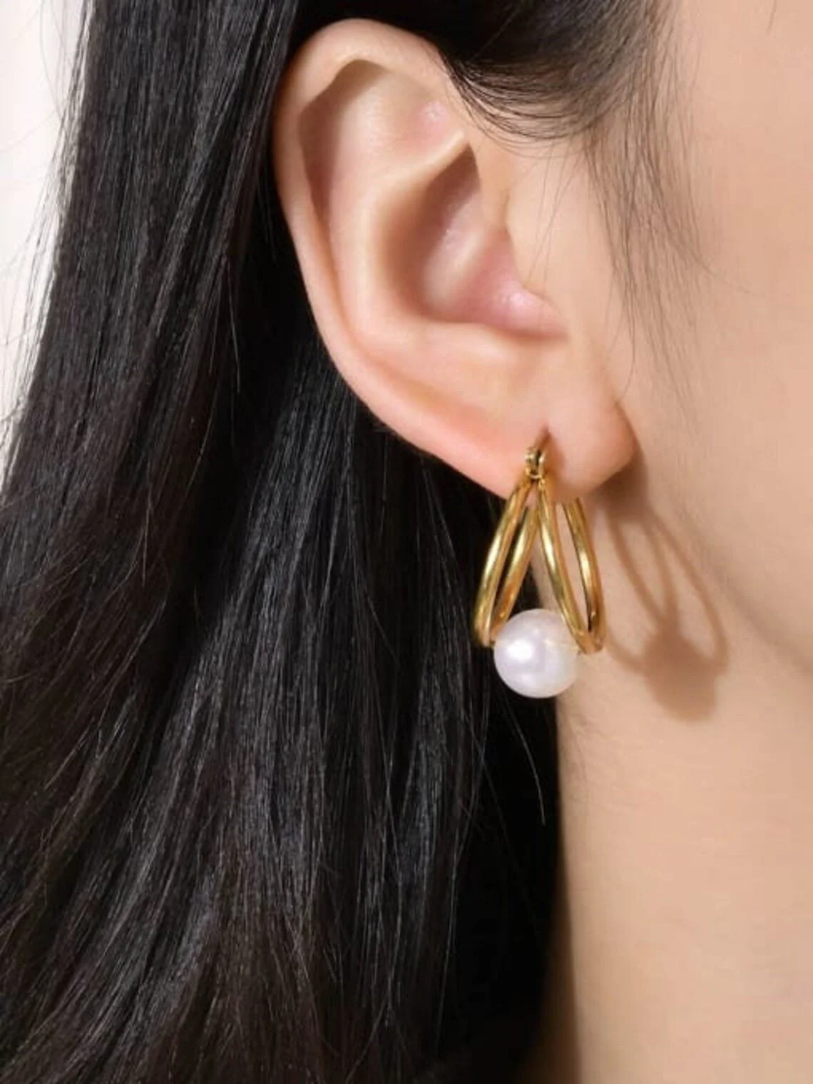 Aretes dorados con una perla colgando, usado en una oreja de una mujer con cabello oscuro.