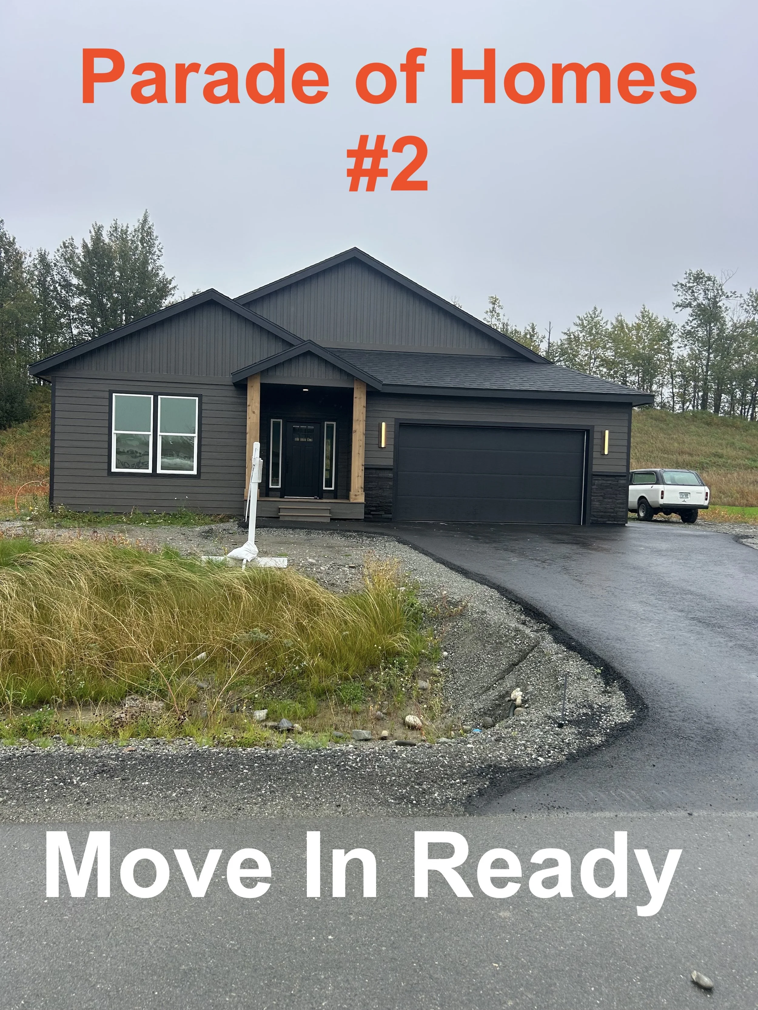 New black house with front entrance, two windows, attached garage, gravel driveway, and a white truck parked on the side, with text overlay indicating 'Parade of Homes #2' and 'Move In Ready'.