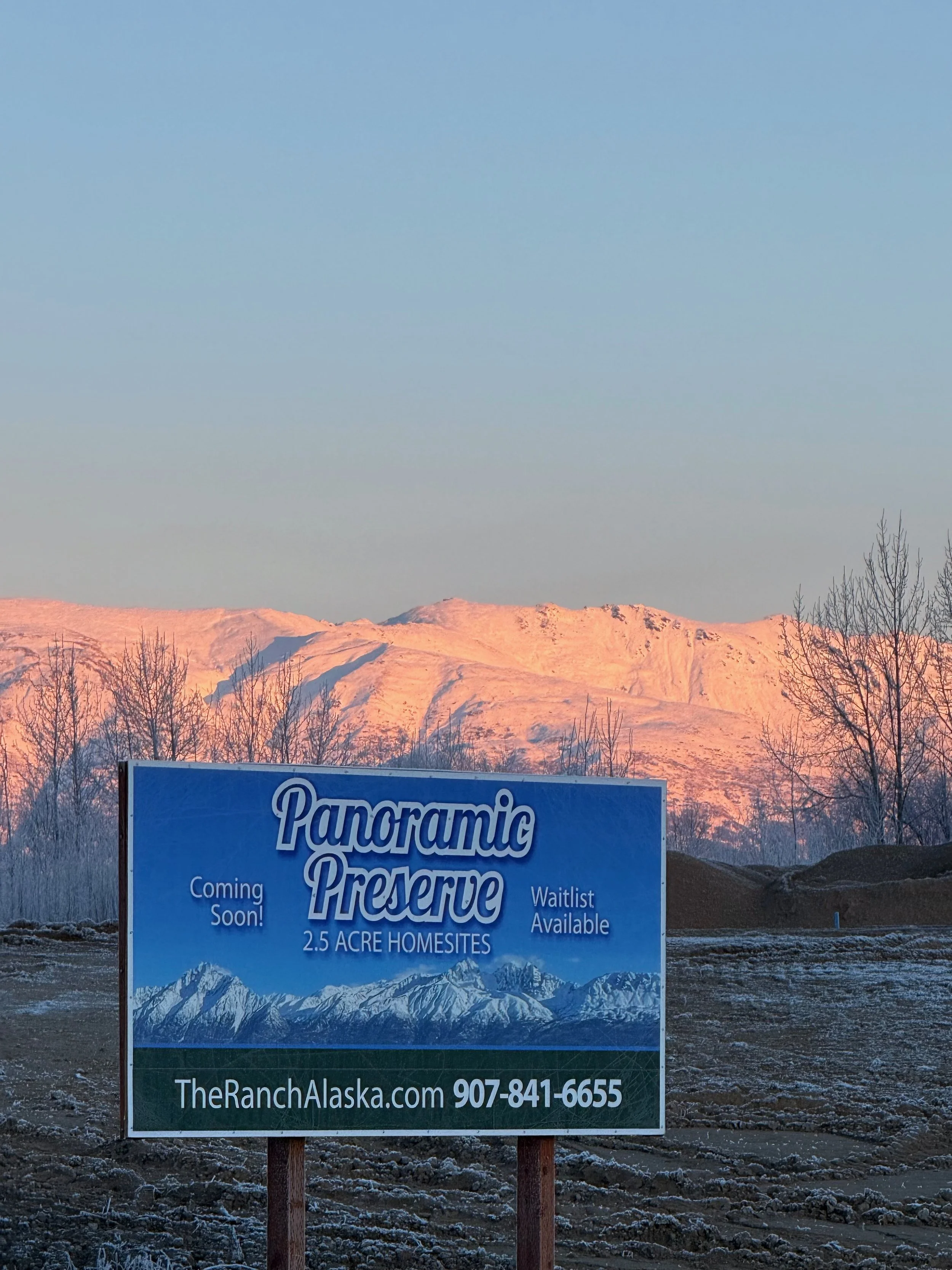 A sign advertising Panoramic Preserve with snow-capped mountains in the background at dawn or dusk, and a landscape with some trees and barren ground.