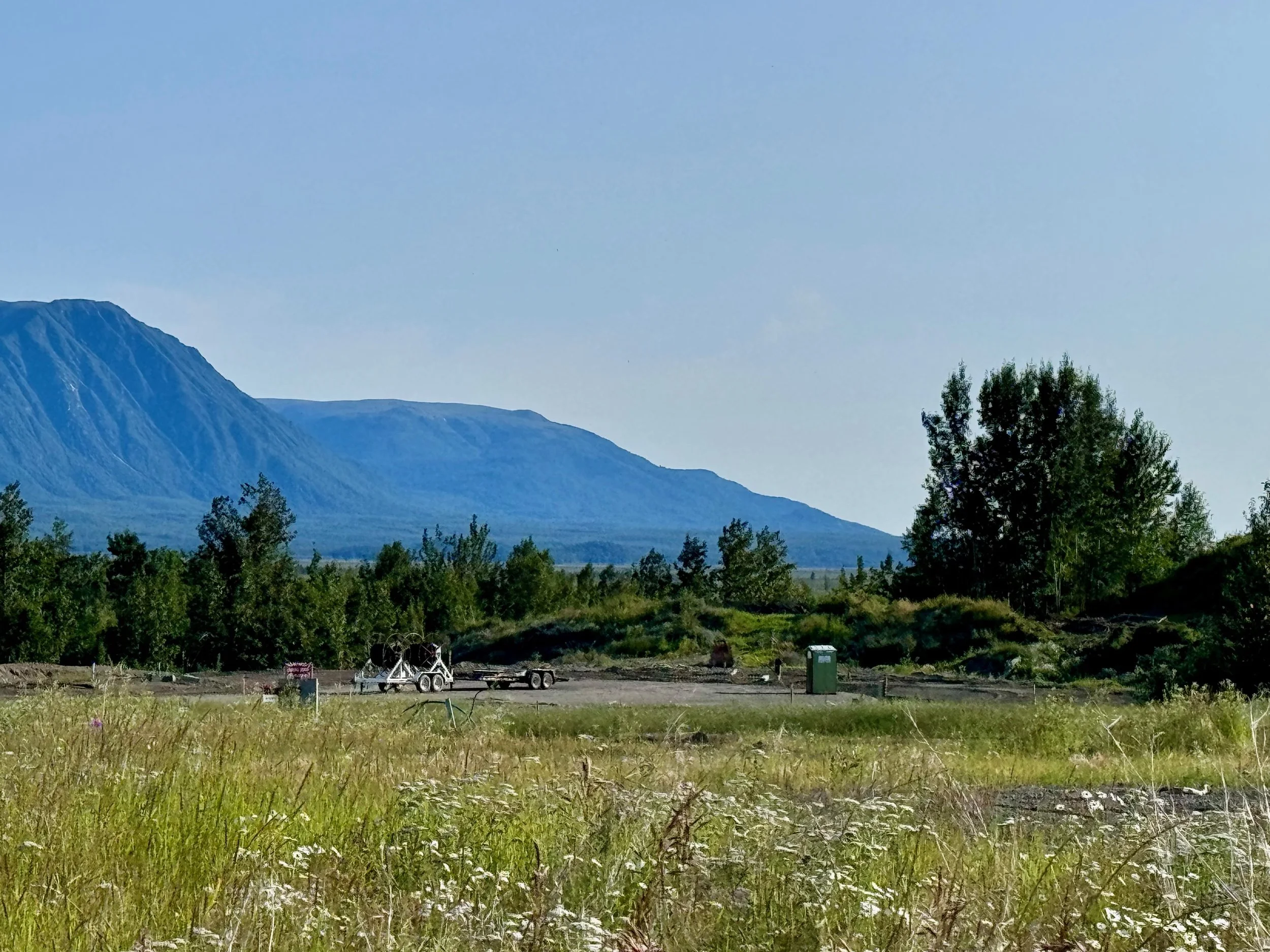 A scenic landscape with a mountain in the background, green trees, and a grassy field in the foreground. There are some construction or maintenance vehicles and equipment on the ground.