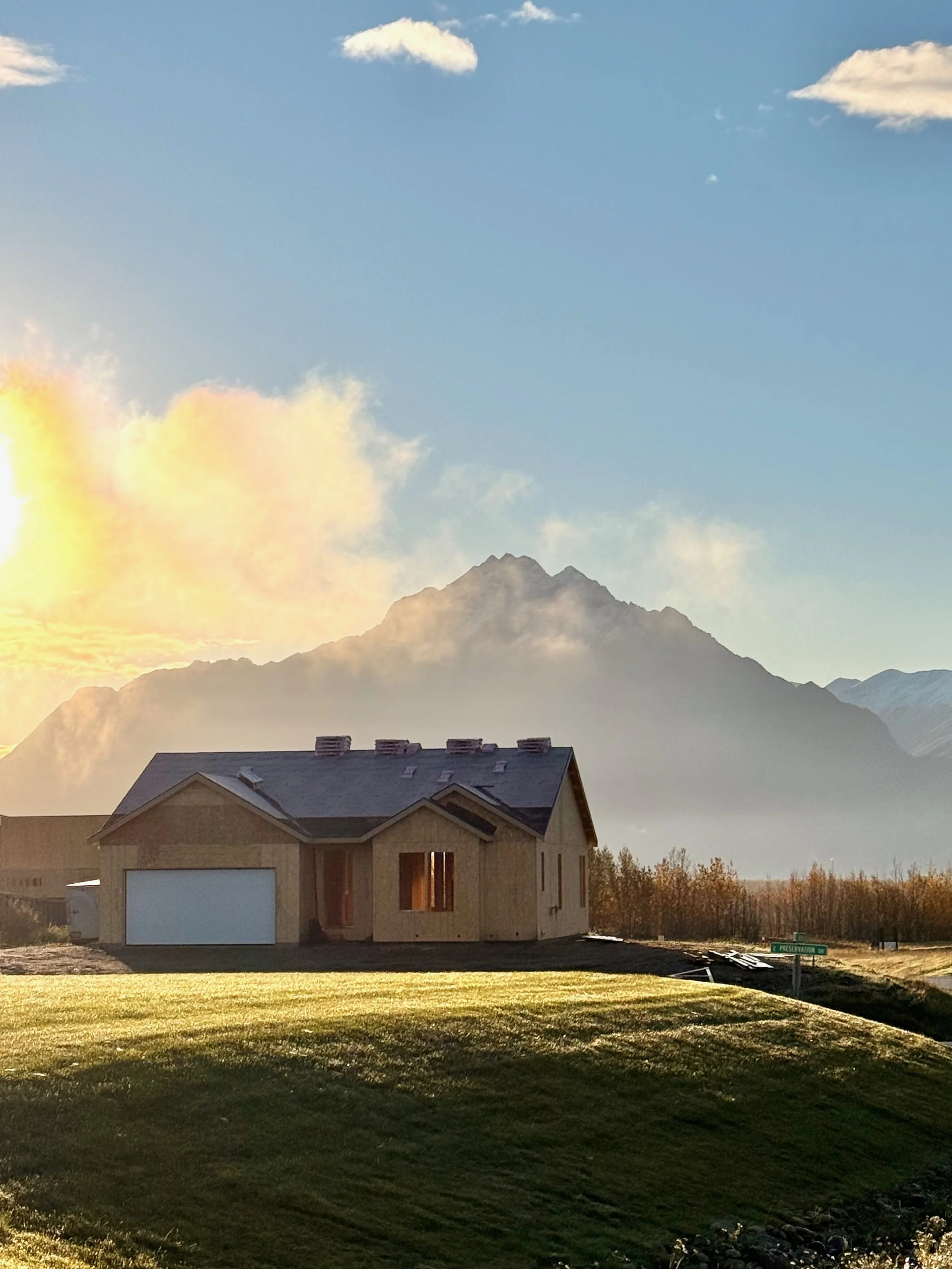A house under construction with a mountainous landscape in the background, under a partly cloudy sky with sunlight.