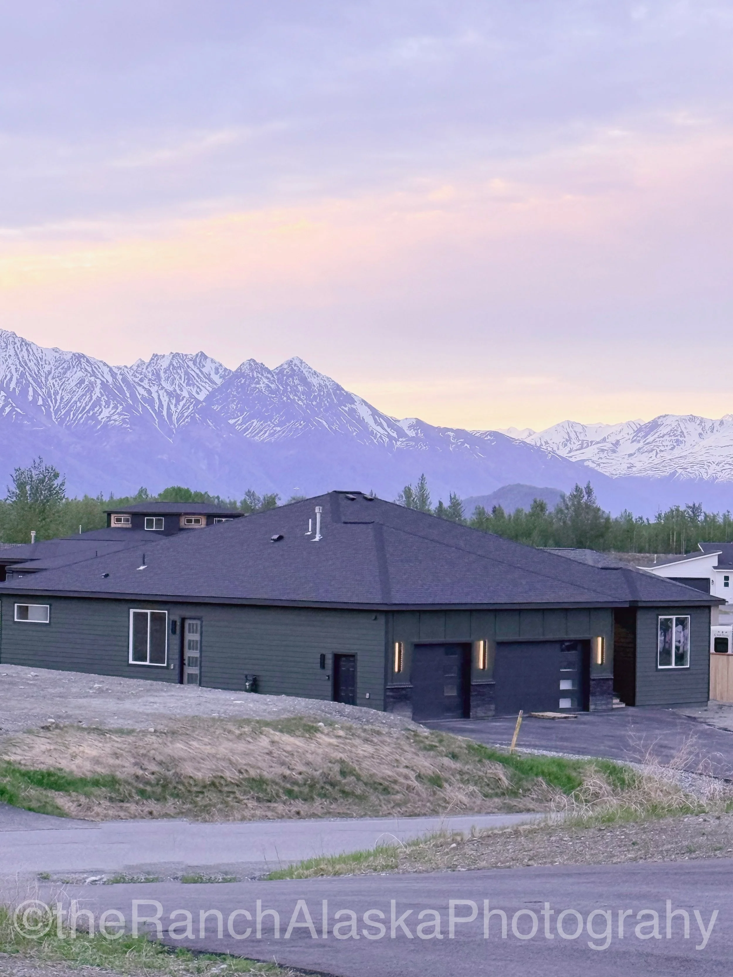 New house under construction with a two-car garage, beige siding, and a single window, on a dirt lot with a mountain range in the background.