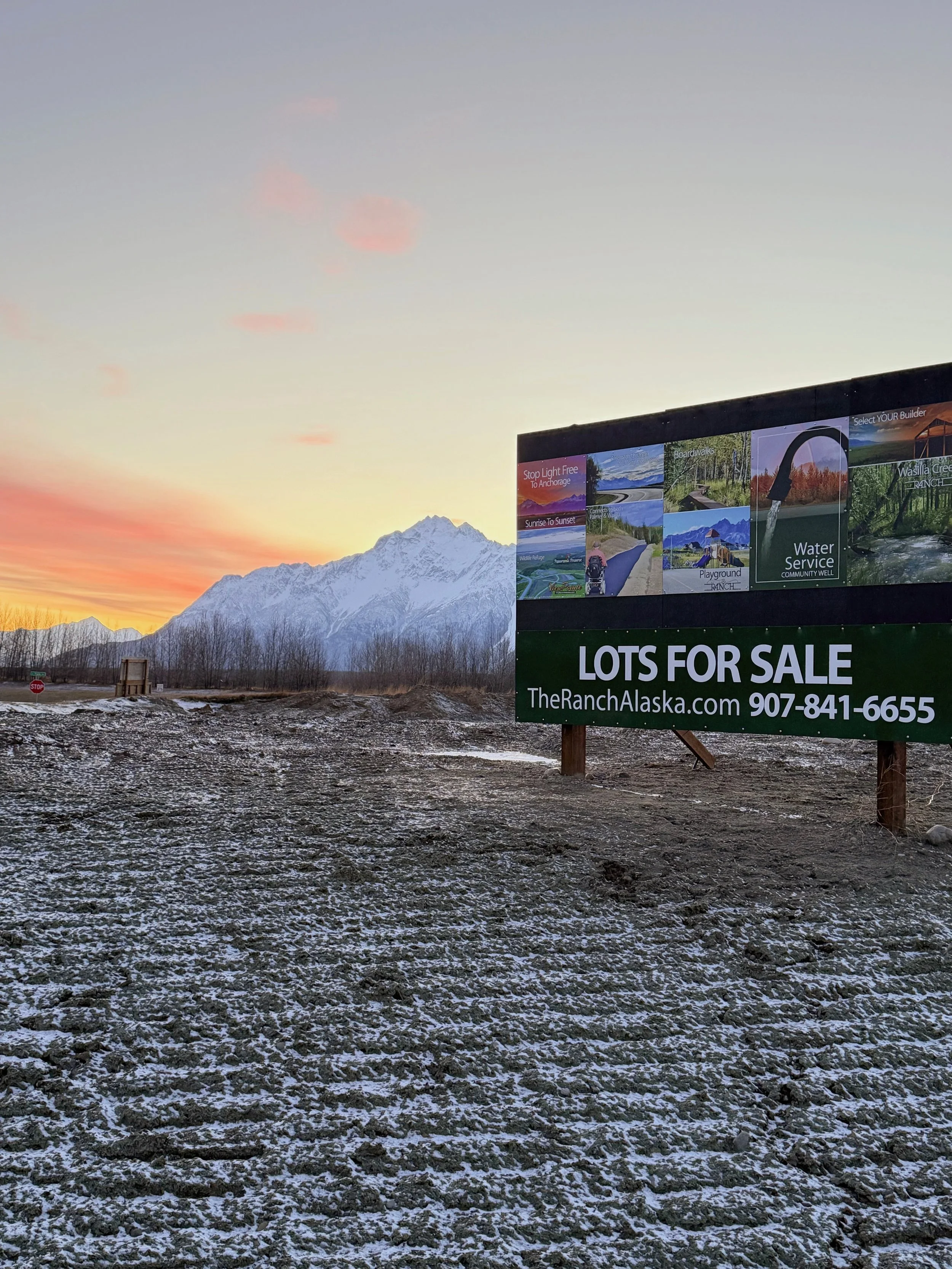 A large outdoor sign advertising lots for sale at TheRanchAlaska.com with a phone number, set against a backdrop of snow-capped mountains during sunset or sunrise, with a partly cloudy sky and a dirt lot in the foreground.