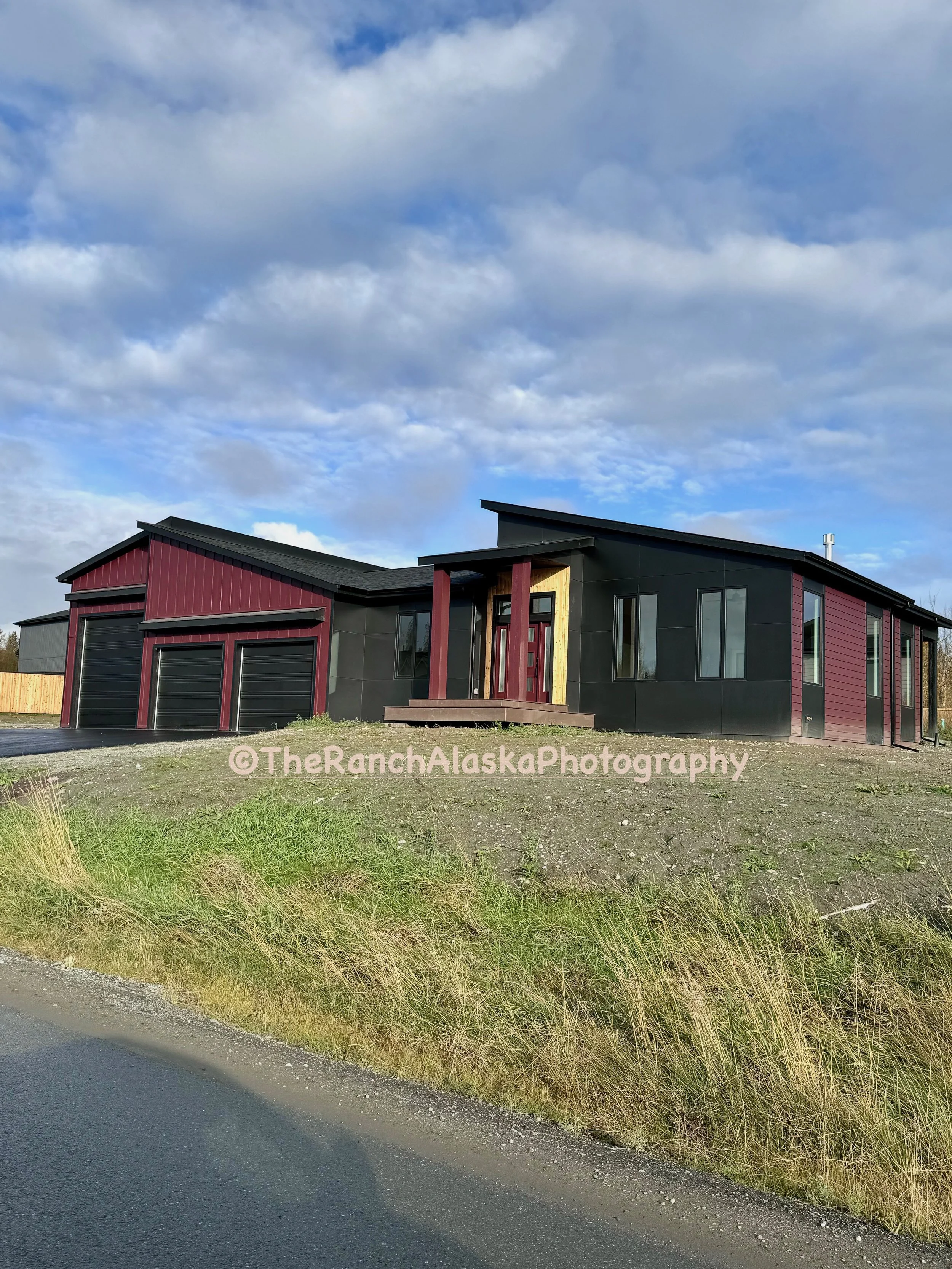 New modern house on a grassy hill with black and red exterior siding, multiple large windows, and a sloped roof, under a partly cloudy sky.