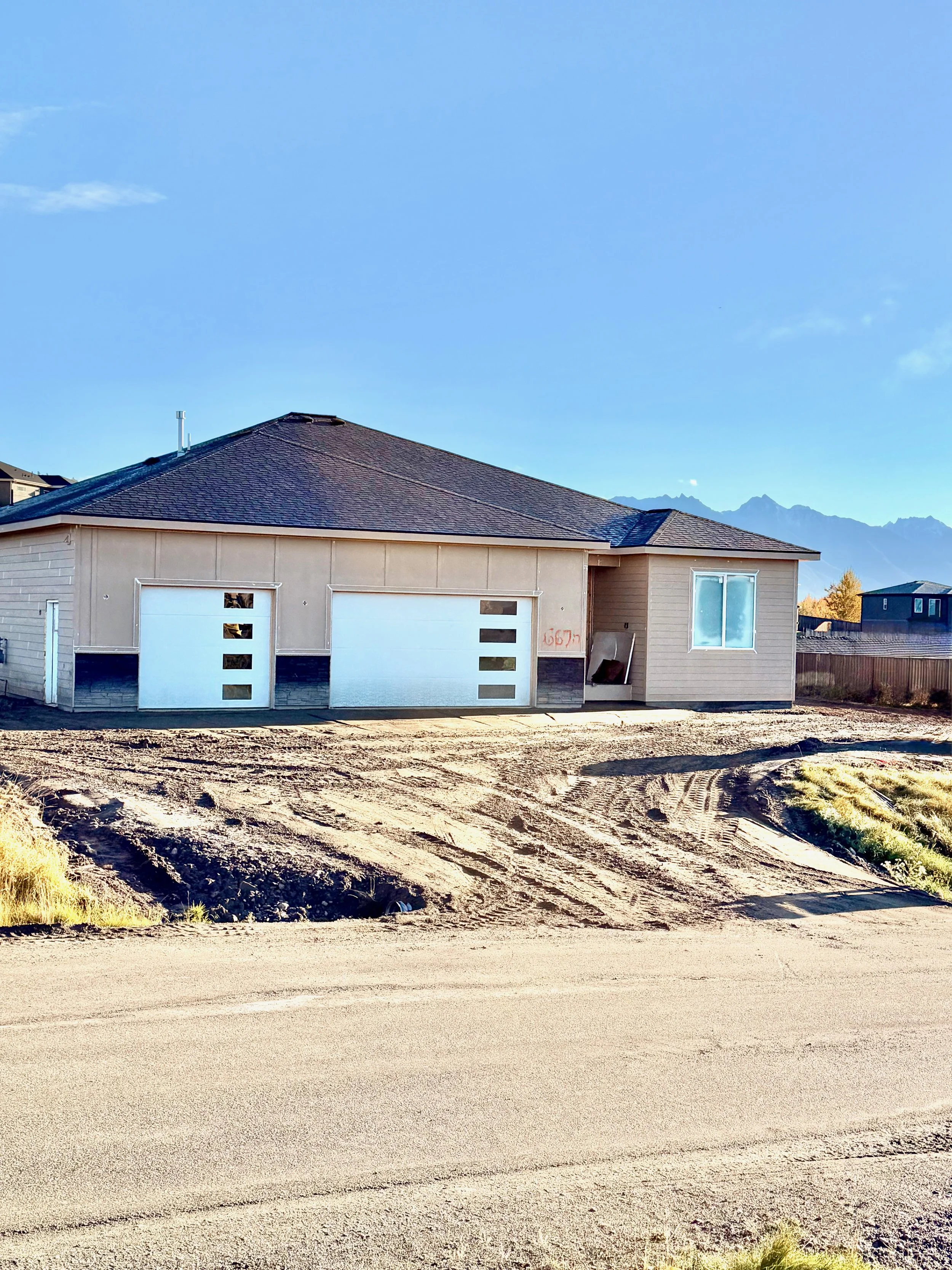 New house under construction with a two-car garage, beige siding, and a single window, on a dirt lot with a mountain range in the background.