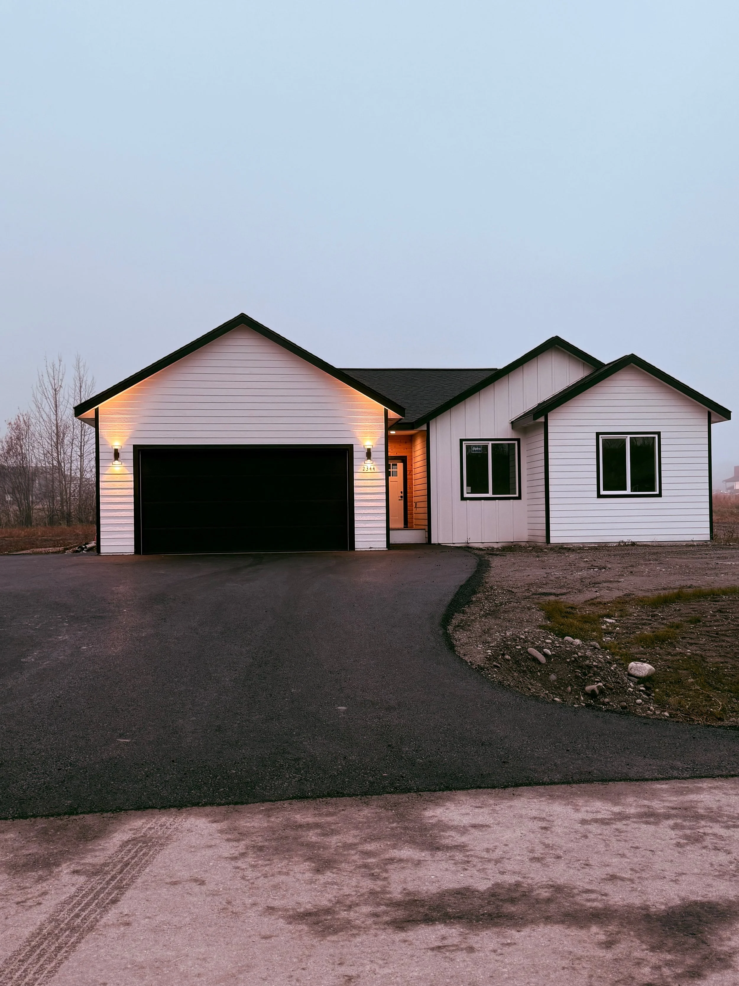 A beige house under construction with a white garage door, situated on a gravel lot, with a backdrop of a blue sky and some trees, and a sign in bold red text that reads 'Ready Soon!! Still time to pick Paint Counter Tops Carpet.'