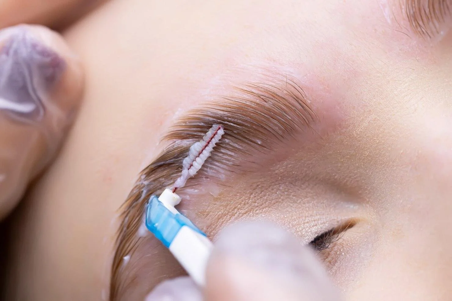 Close-up of a person receiving eyelash extension with a technician applying adhesive to natural lashes.