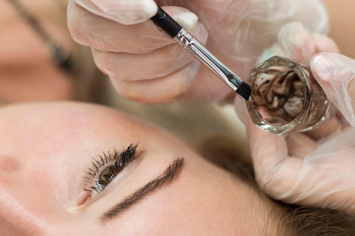 Beautician applying eyebrow tattoo with a small brush and ink in a glass container to a woman lying down with closed eyes.
