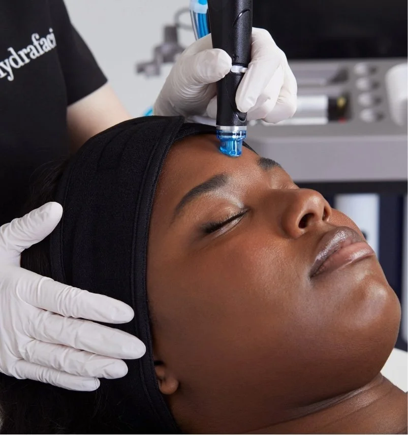 A woman receives a facial treatment with a handheld device at a skincare clinic, lying with her eyes closed and wearing a black headband.