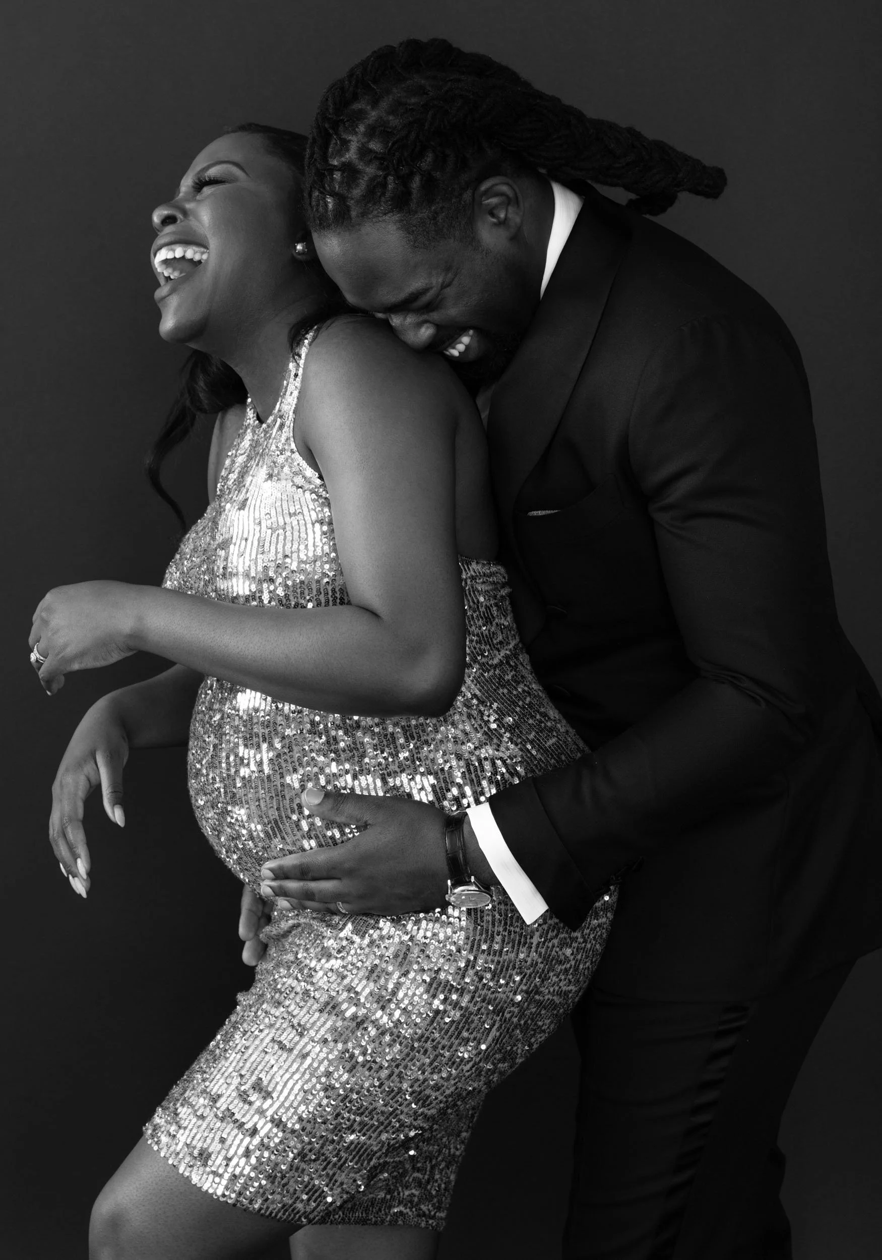 Black-and-white maternity portrait of an expectant couple laughing and embracing during their studio session.