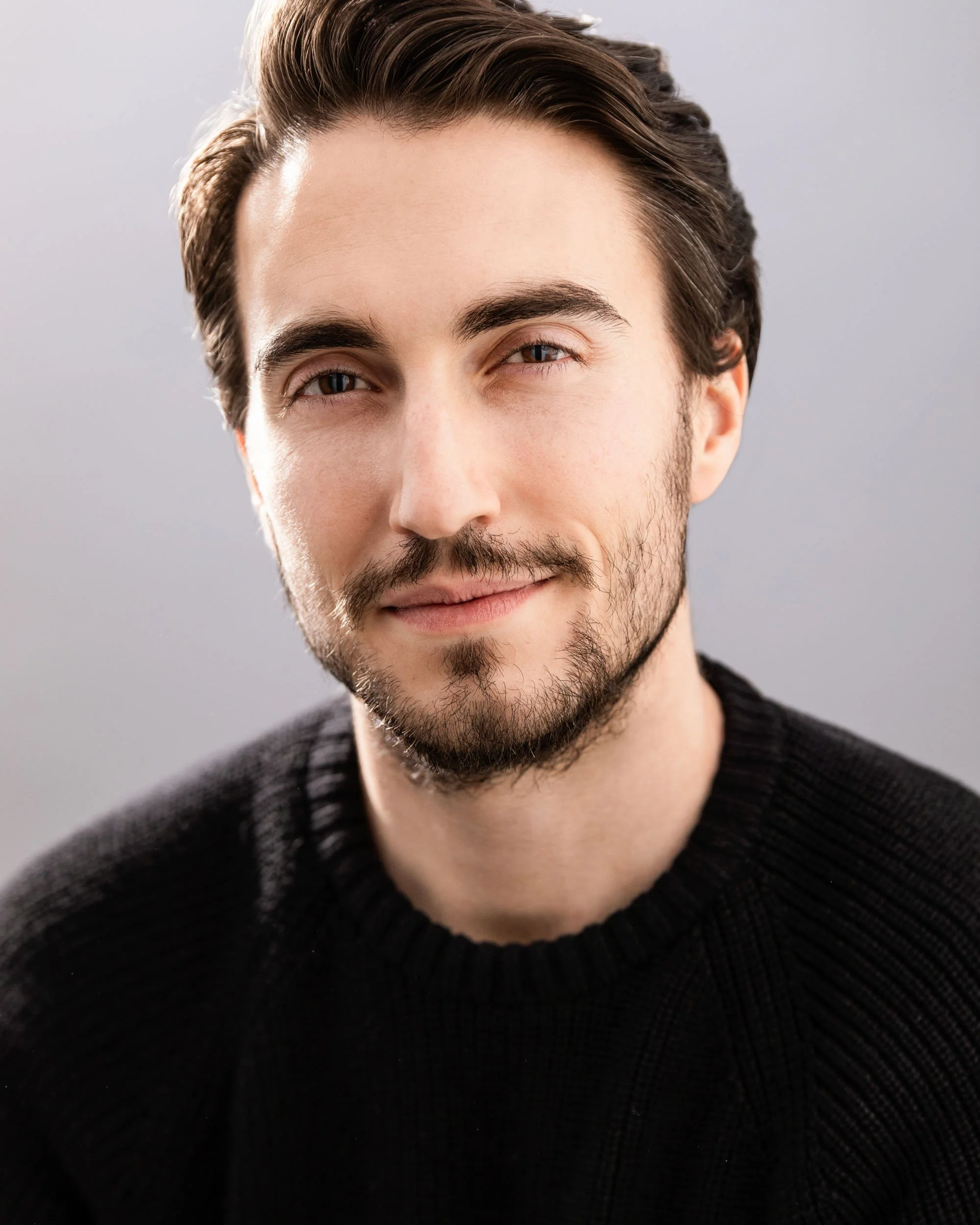 Close professional headshot of a man with dark hair and a short beard wearing a black sweater against a soft gray background.