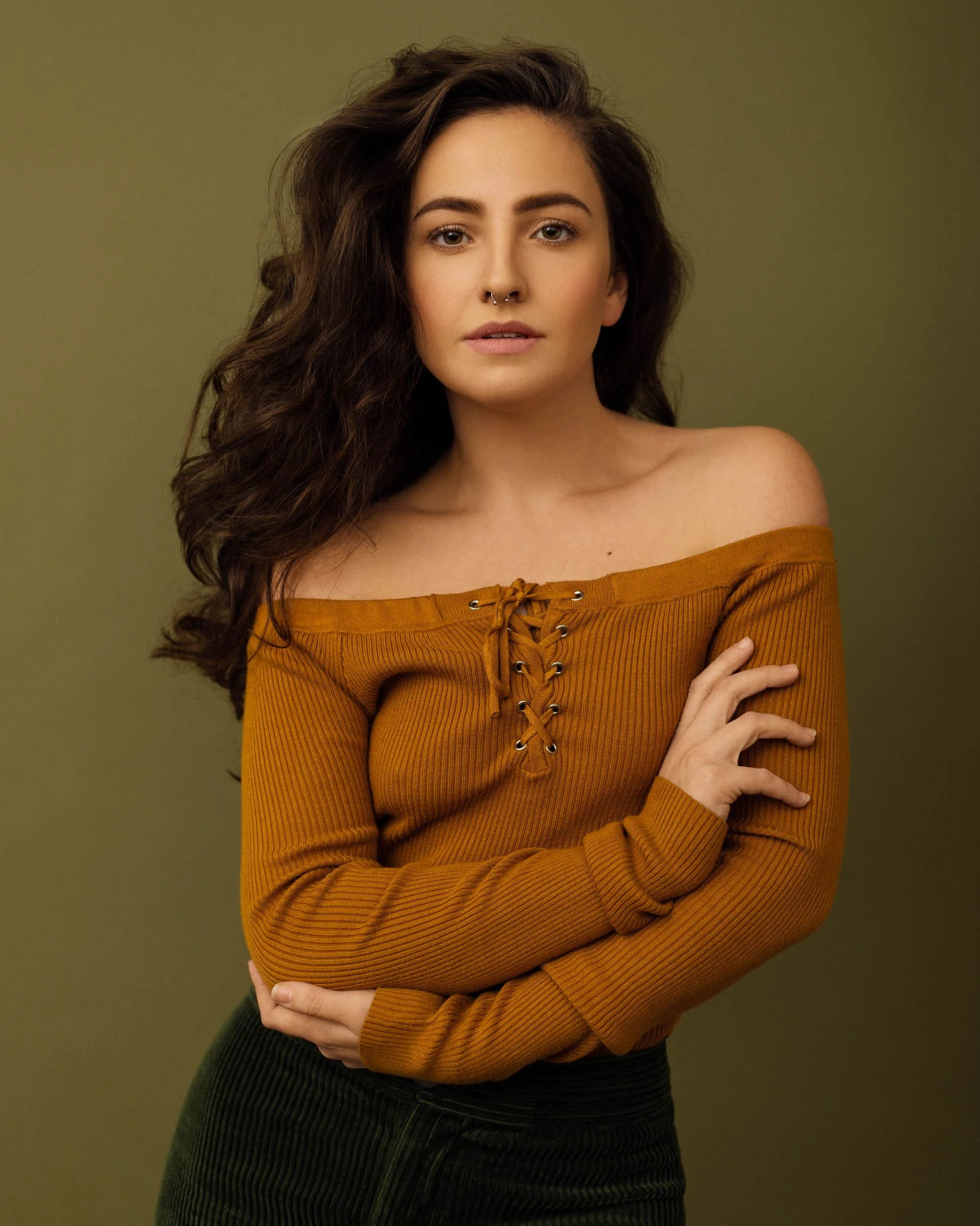 Editorial portrait of a woman with long dark wavy hair wearing a mustard off-the-shoulder sweater against an olive background.
