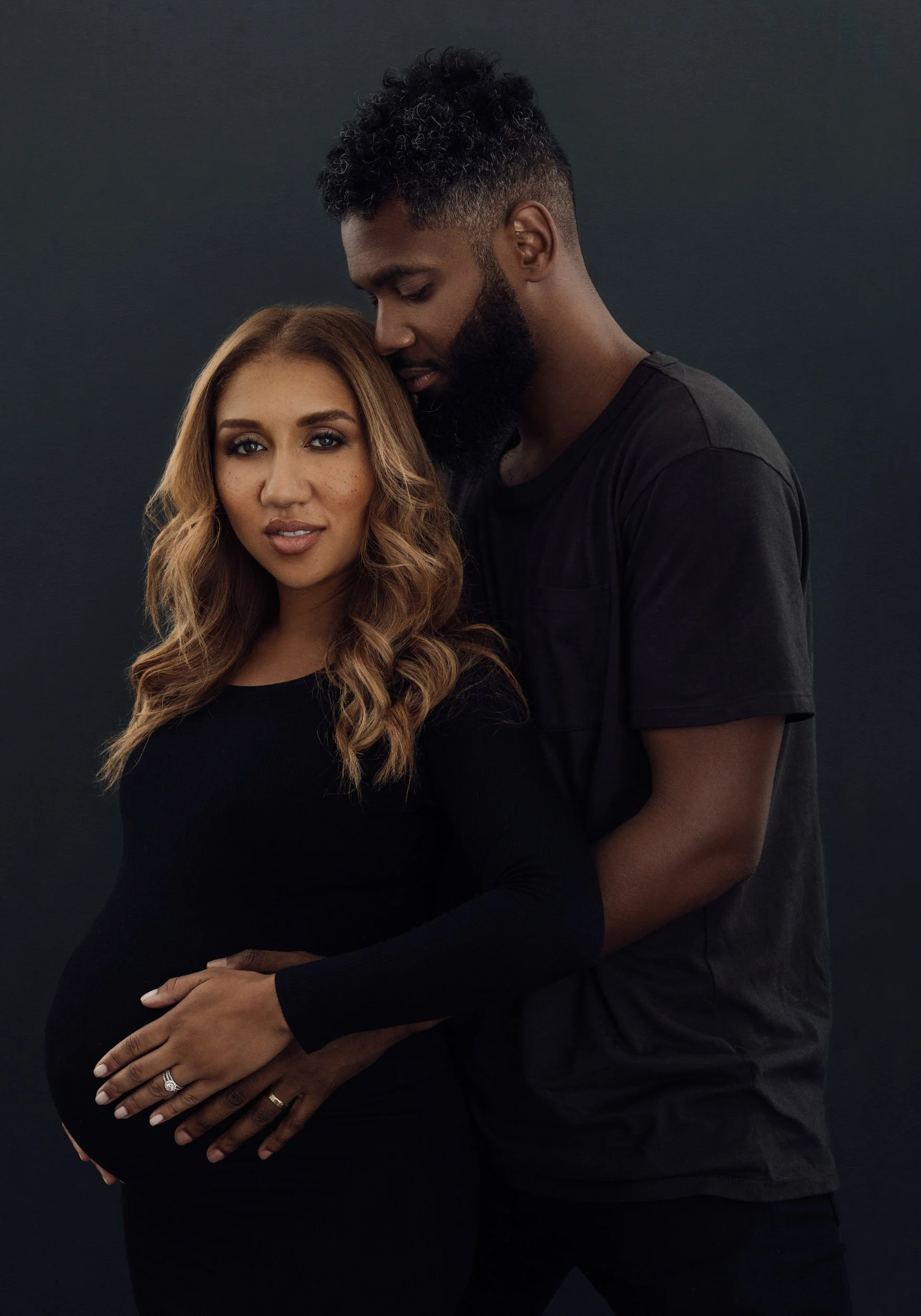 Intimate maternity portrait of an expectant couple standing closely together against a dark studio background.