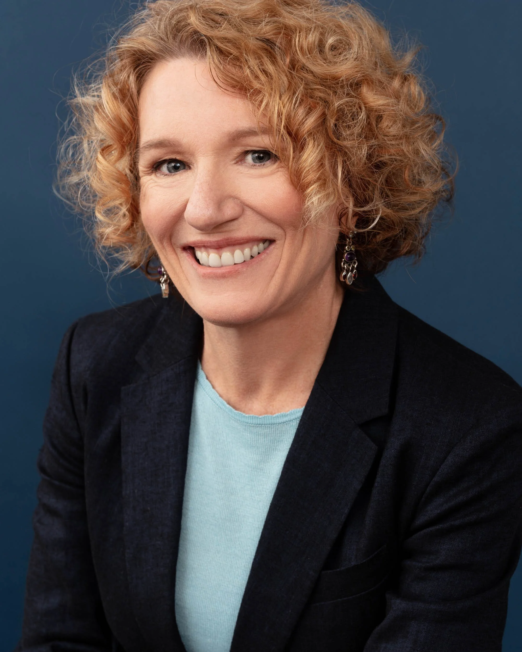 Professional headshot of a smiling woman with short curly red hair wearing a navy blazer and light blue top against a deep blue background.