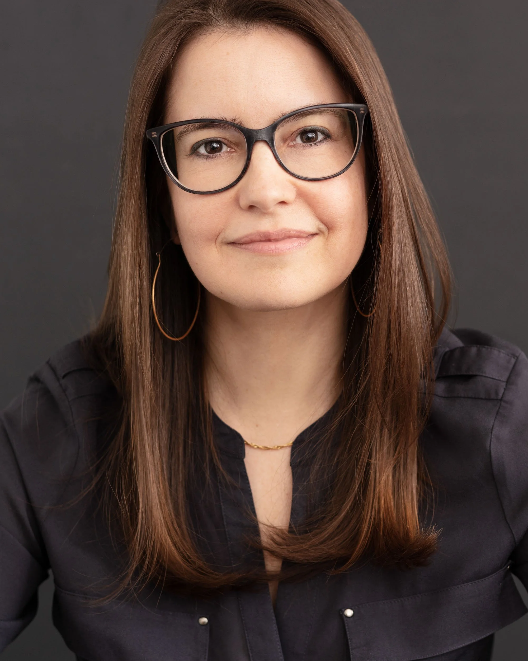 Professional studio headshot of a woman with long brown hair and black glasses wearing a black blouse against a gray background.