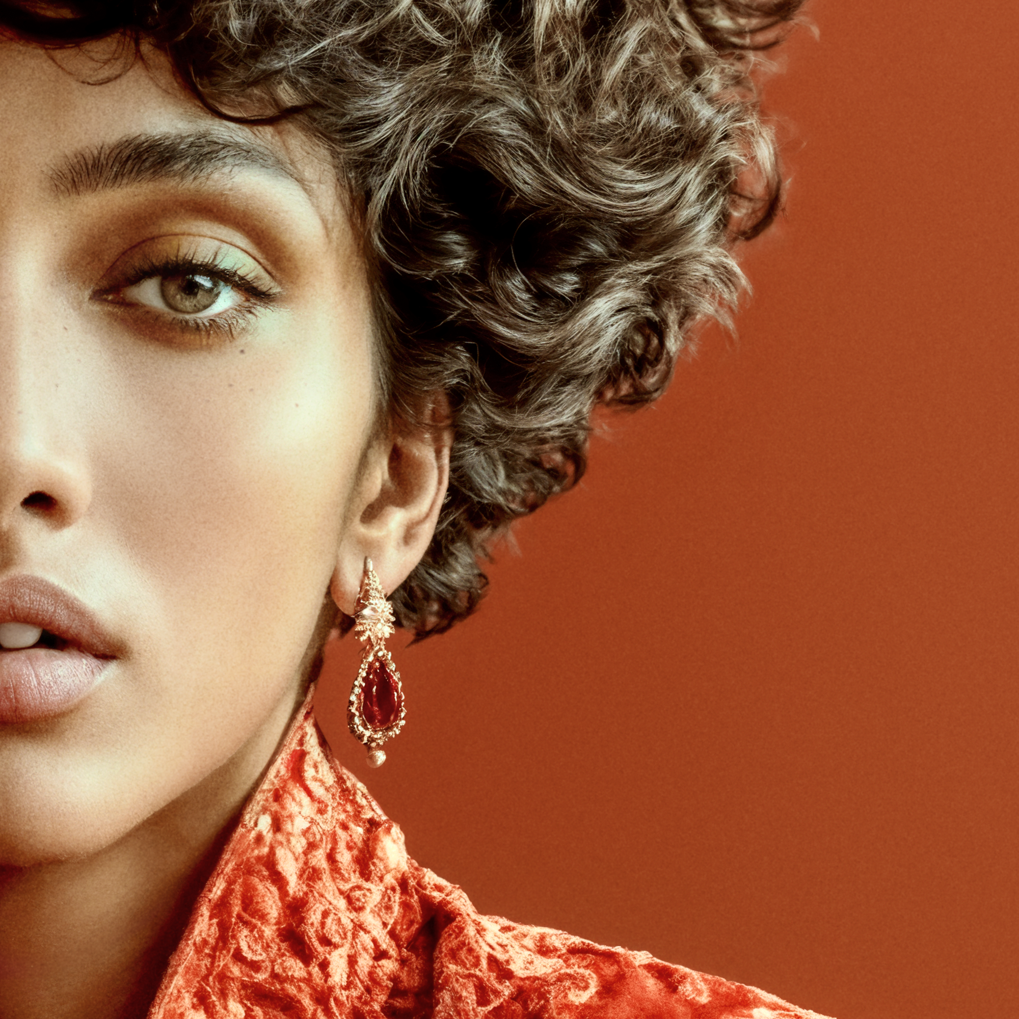 Detail of Kavya wearing a red drop earring beside dark curls and a raised brocade collar.