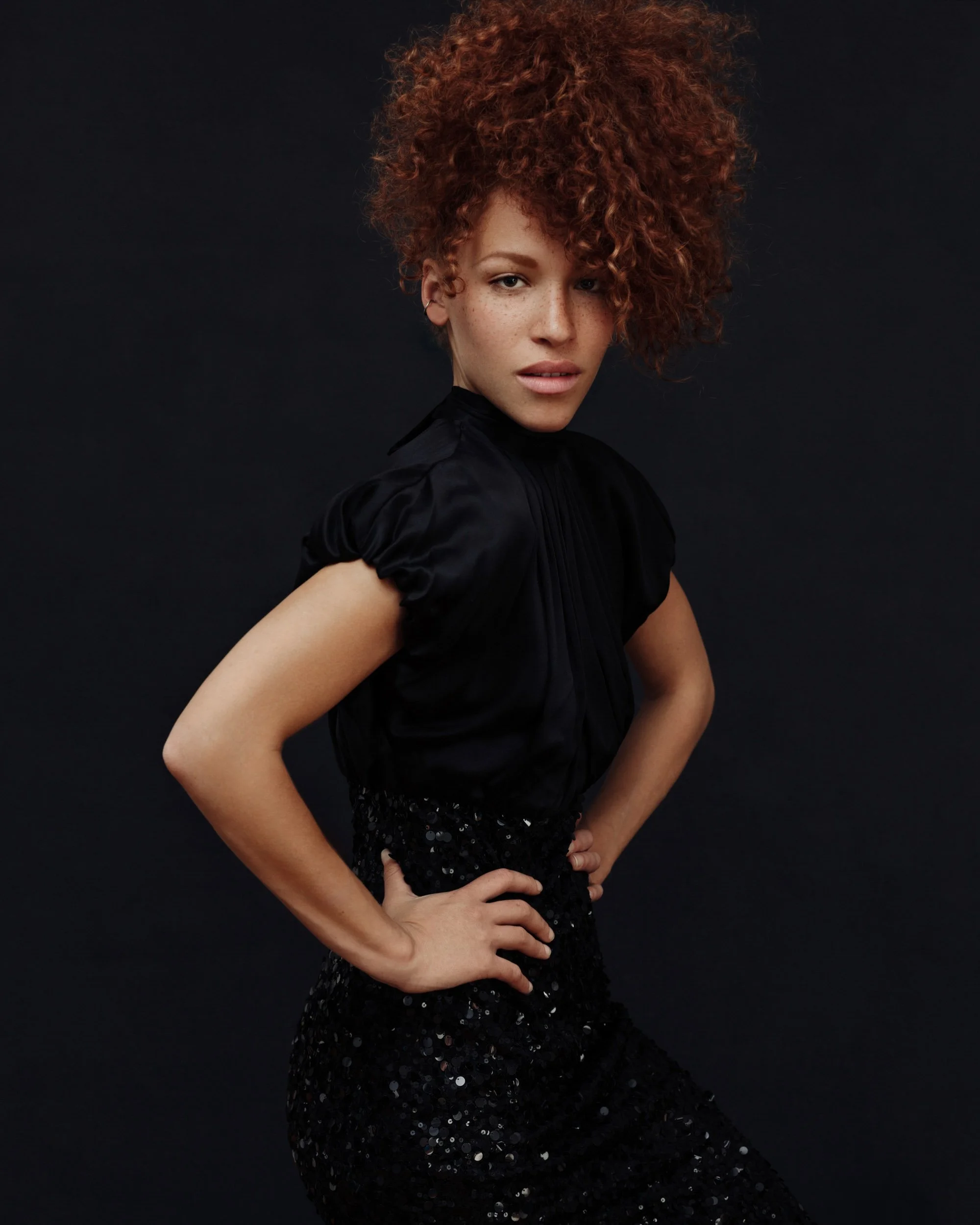 Editorial studio portrait of a woman with voluminous red curls wearing a black dress against a dark background.