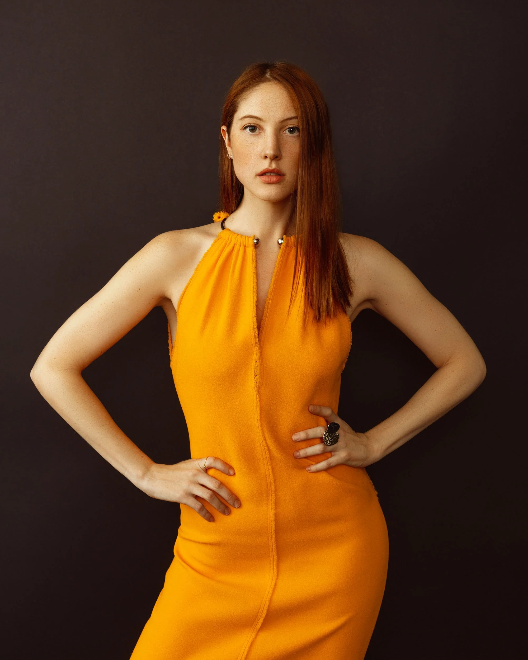 Full-length fashion portrait of a red-haired woman wearing a bright orange dress with her hands on her hips against a dark brown background.