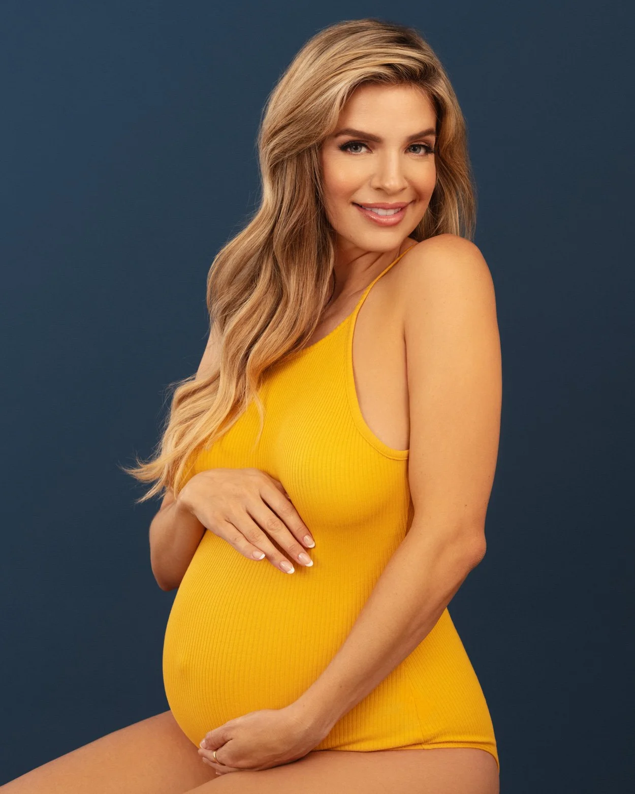 Colorful maternity portrait of a pregnant woman wearing a yellow bodysuit while seated against a deep blue background.