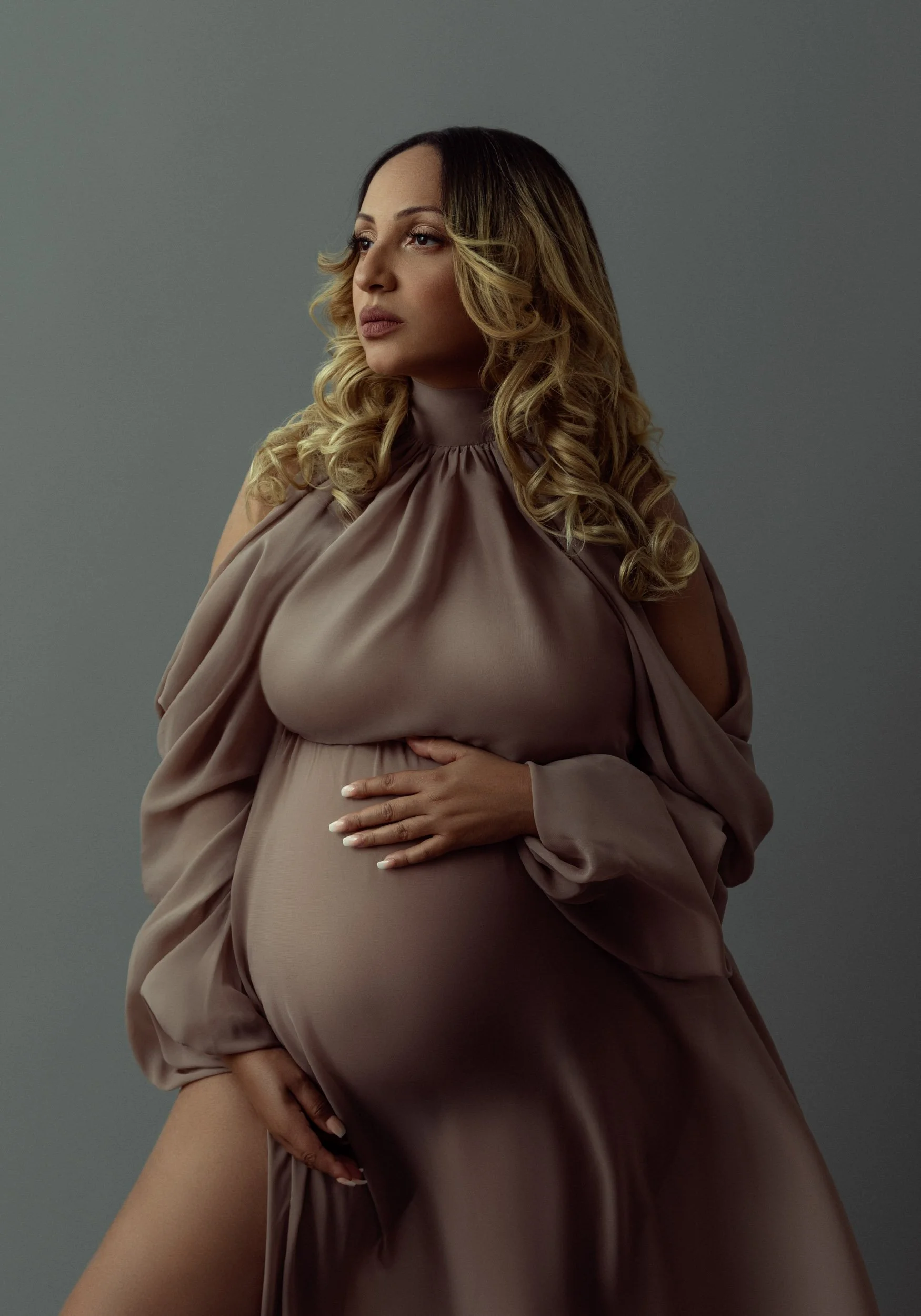 Studio maternity portrait of a pregnant woman wearing a flowing taupe dress against a soft gray background.