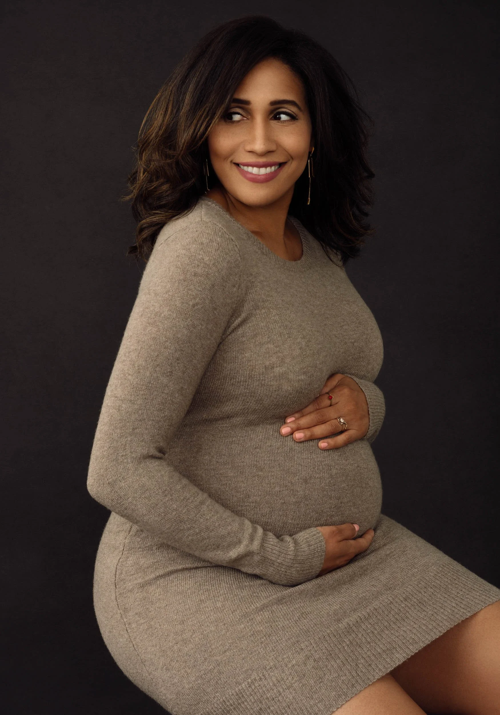Seated studio maternity portrait of a pregnant woman wearing a fitted taupe knit dress against a dark neutral background.