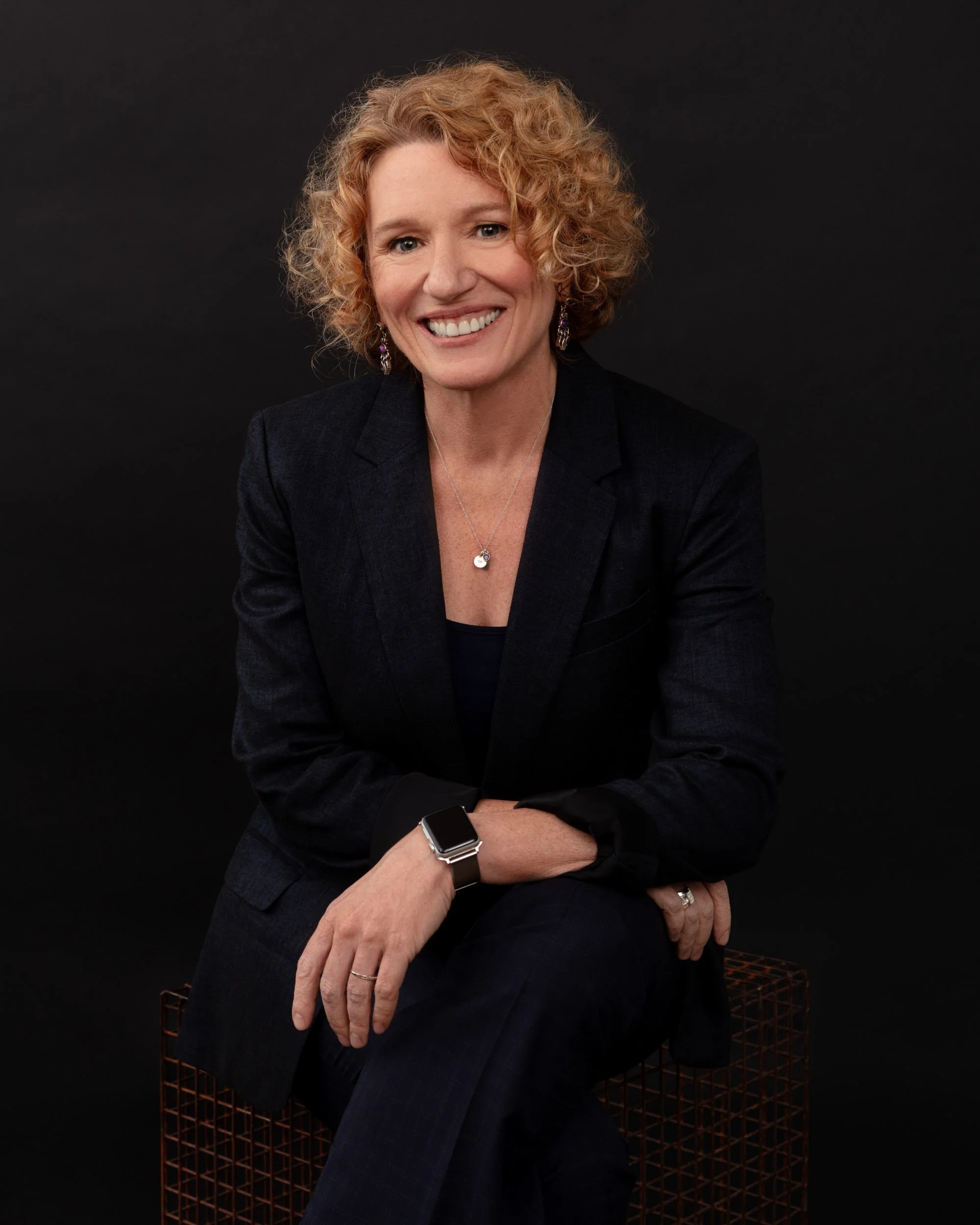 Seated studio portrait of a smiling woman with short curly blonde hair wearing a navy suit against a dark background.