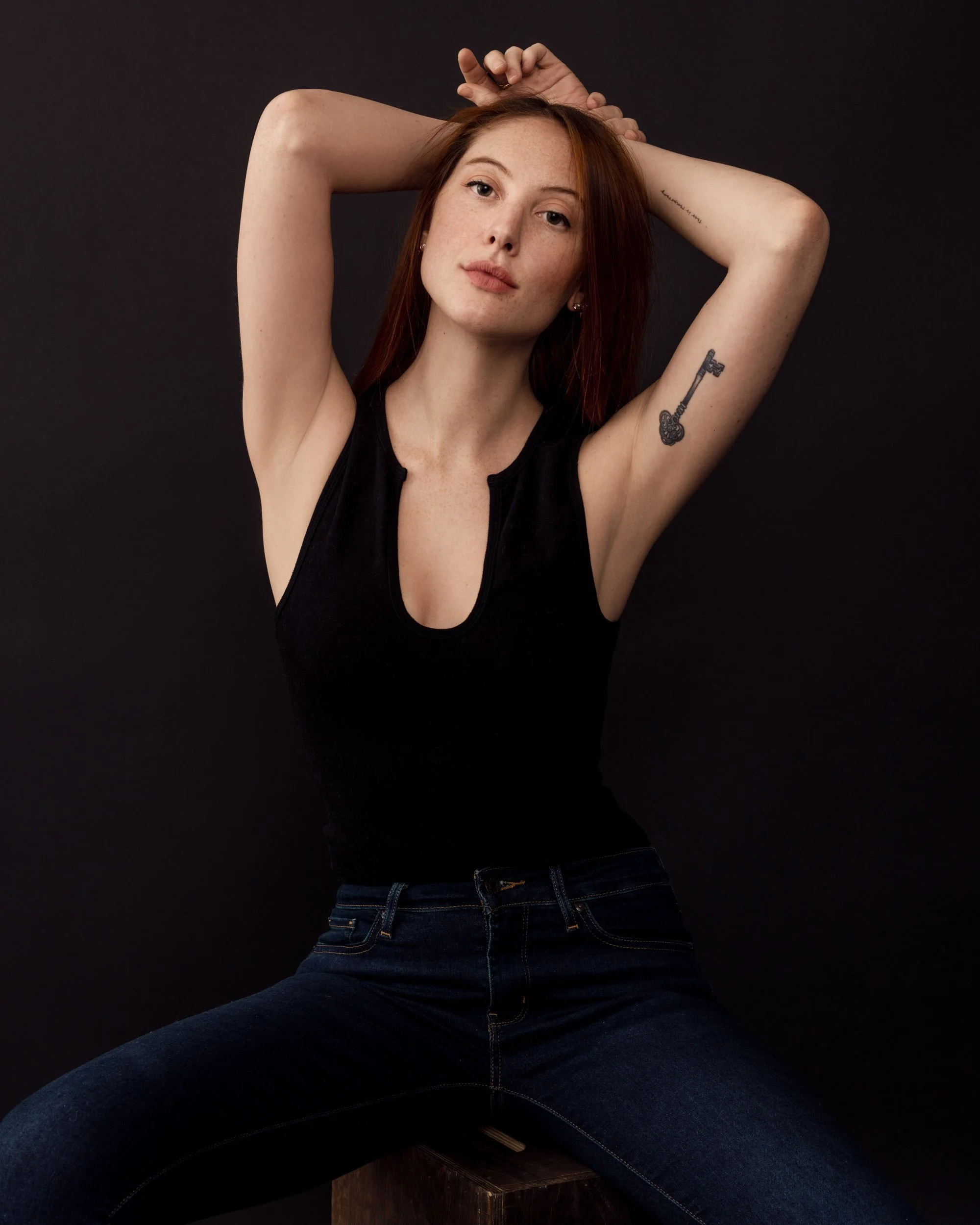 Creative seated portrait of a red-haired woman in a black sleeveless top and jeans posing with both arms raised.