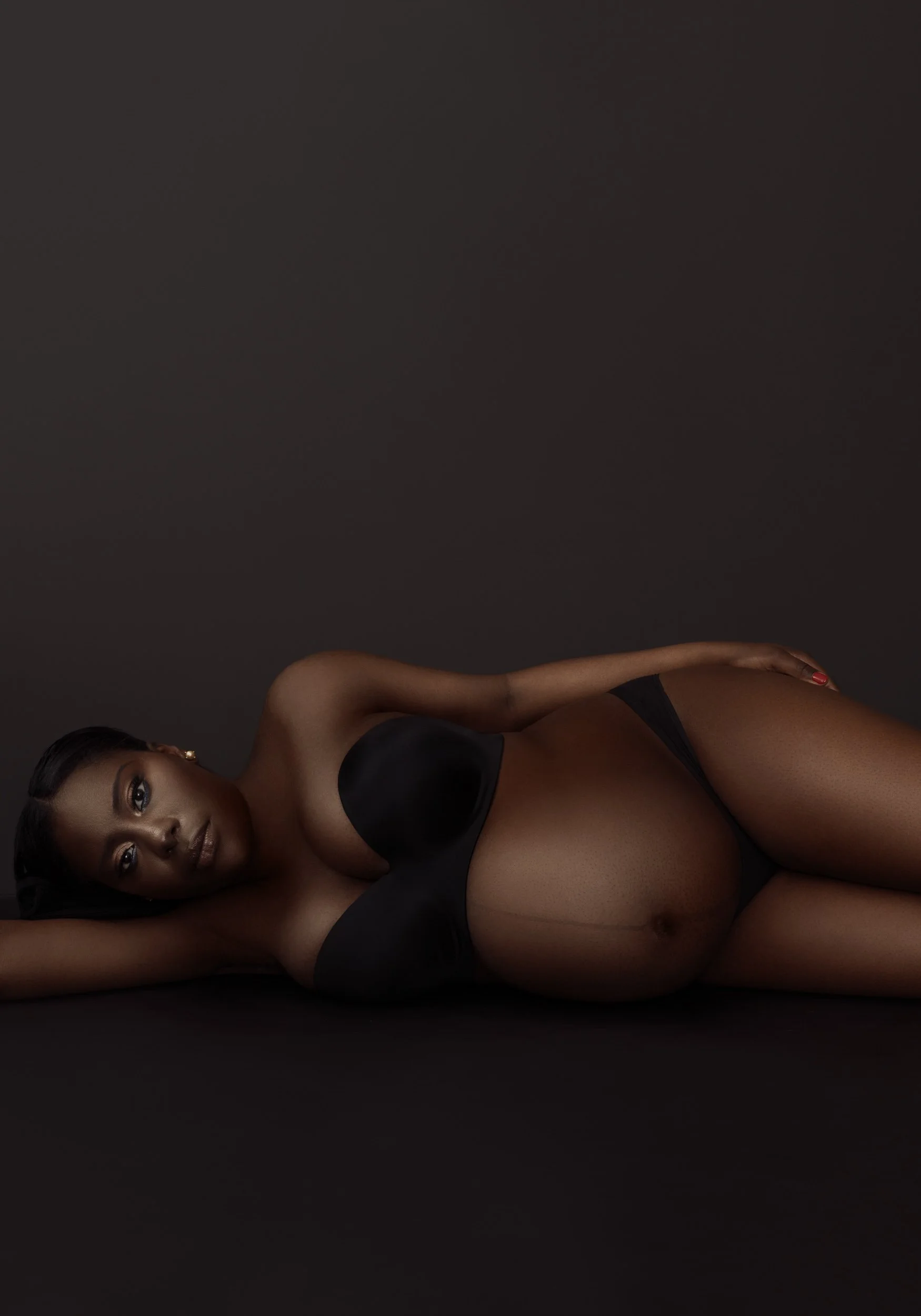 Fine-art maternity portrait of a pregnant woman reclining on her side against a warm brown studio background.