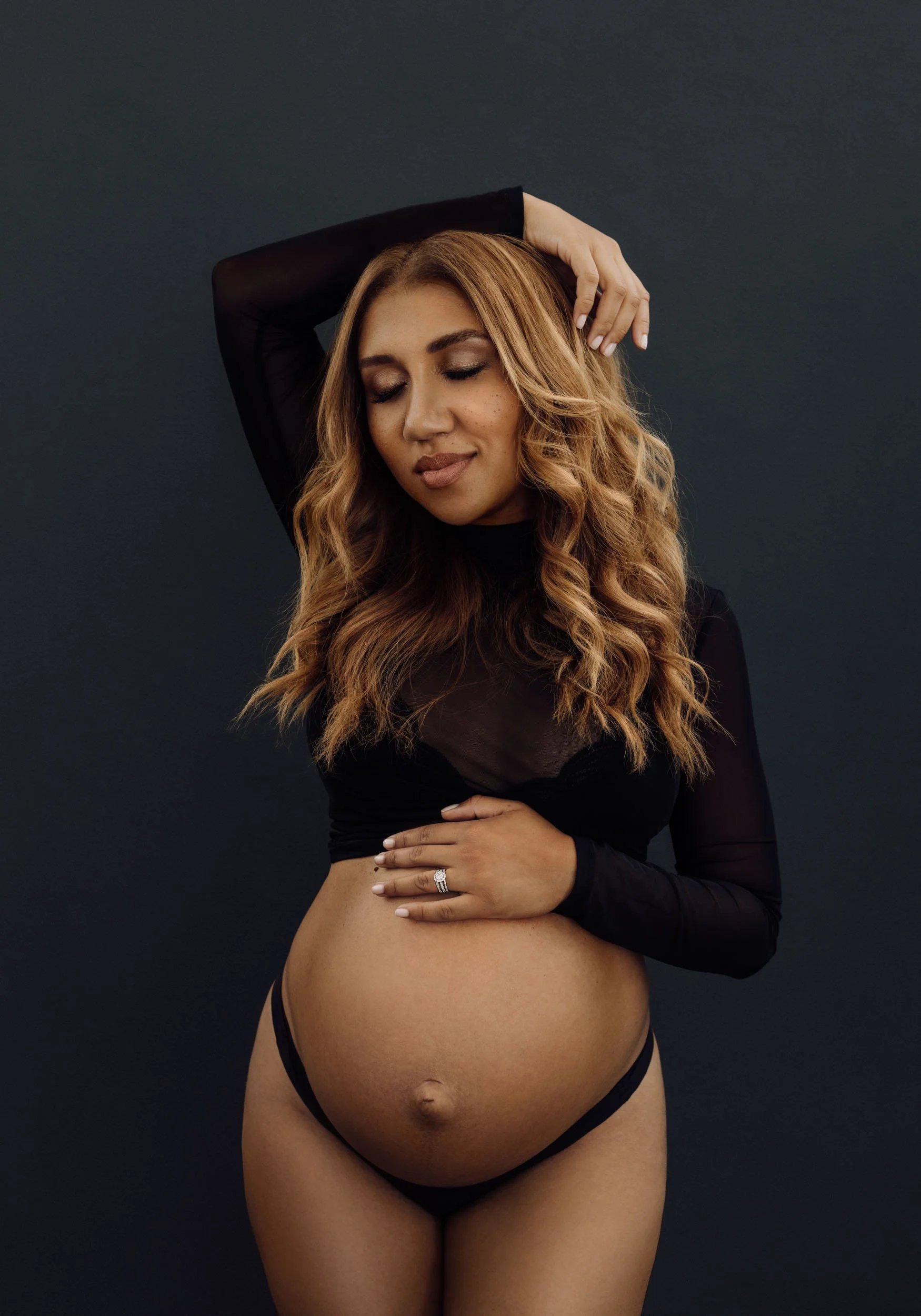 Studio maternity portrait of a pregnant woman in a black bodysuit posing with one arm raised against a dark gray background.