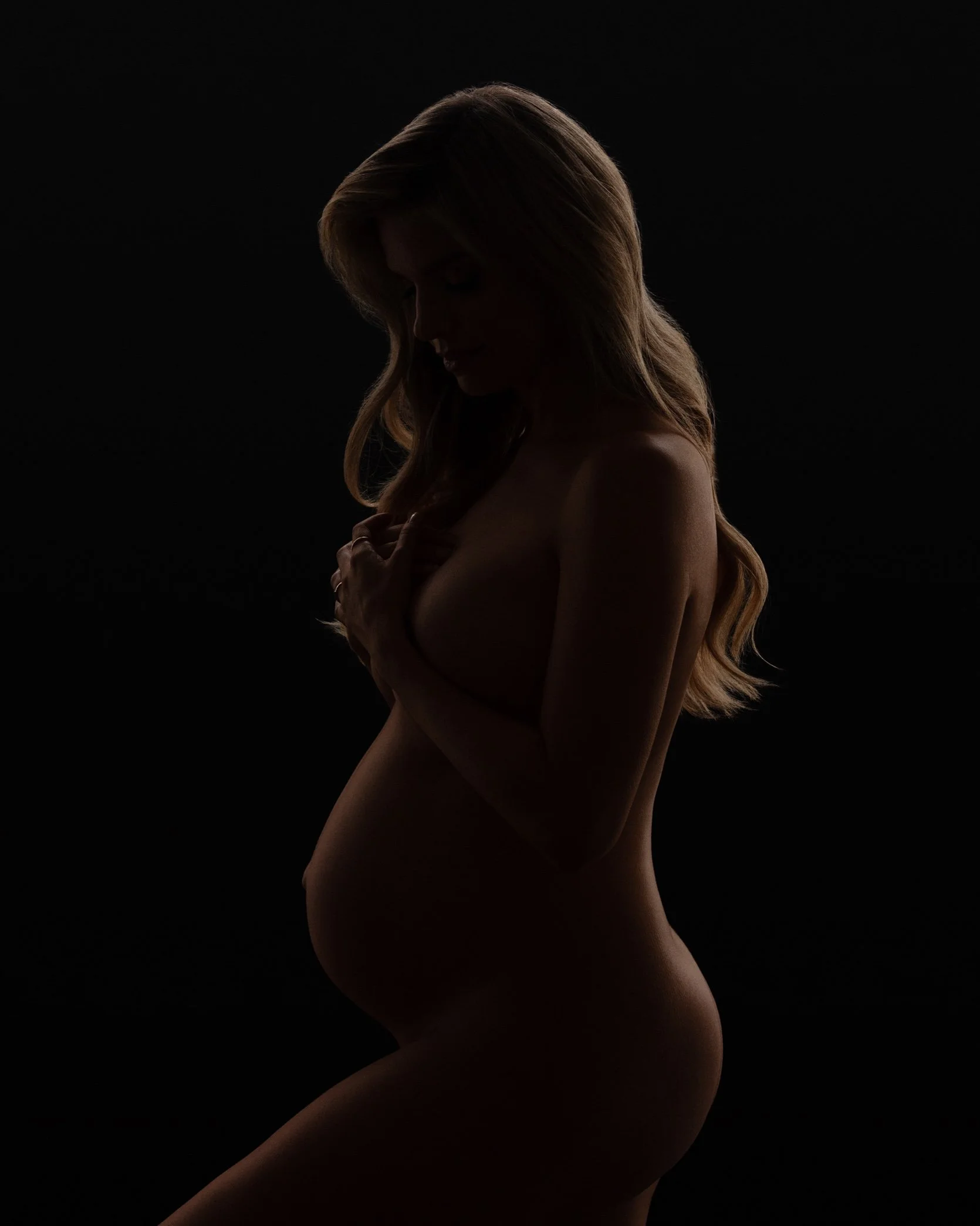 Low-key silhouette maternity portrait showing the profile of a pregnant woman against a nearly black background.