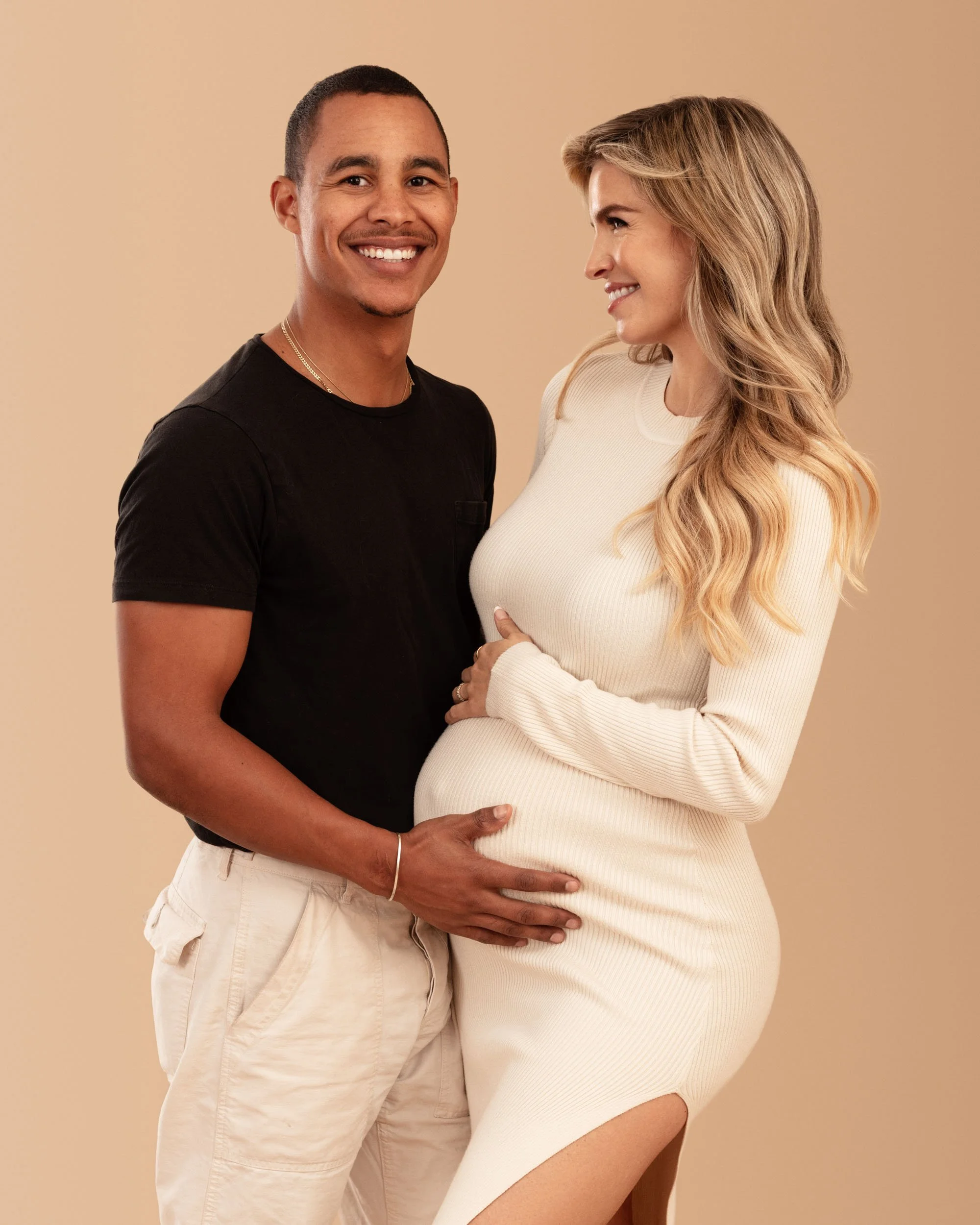 Maternity portrait of an expectant couple wearing coordinated neutral clothing and smiling together against a warm studio background.