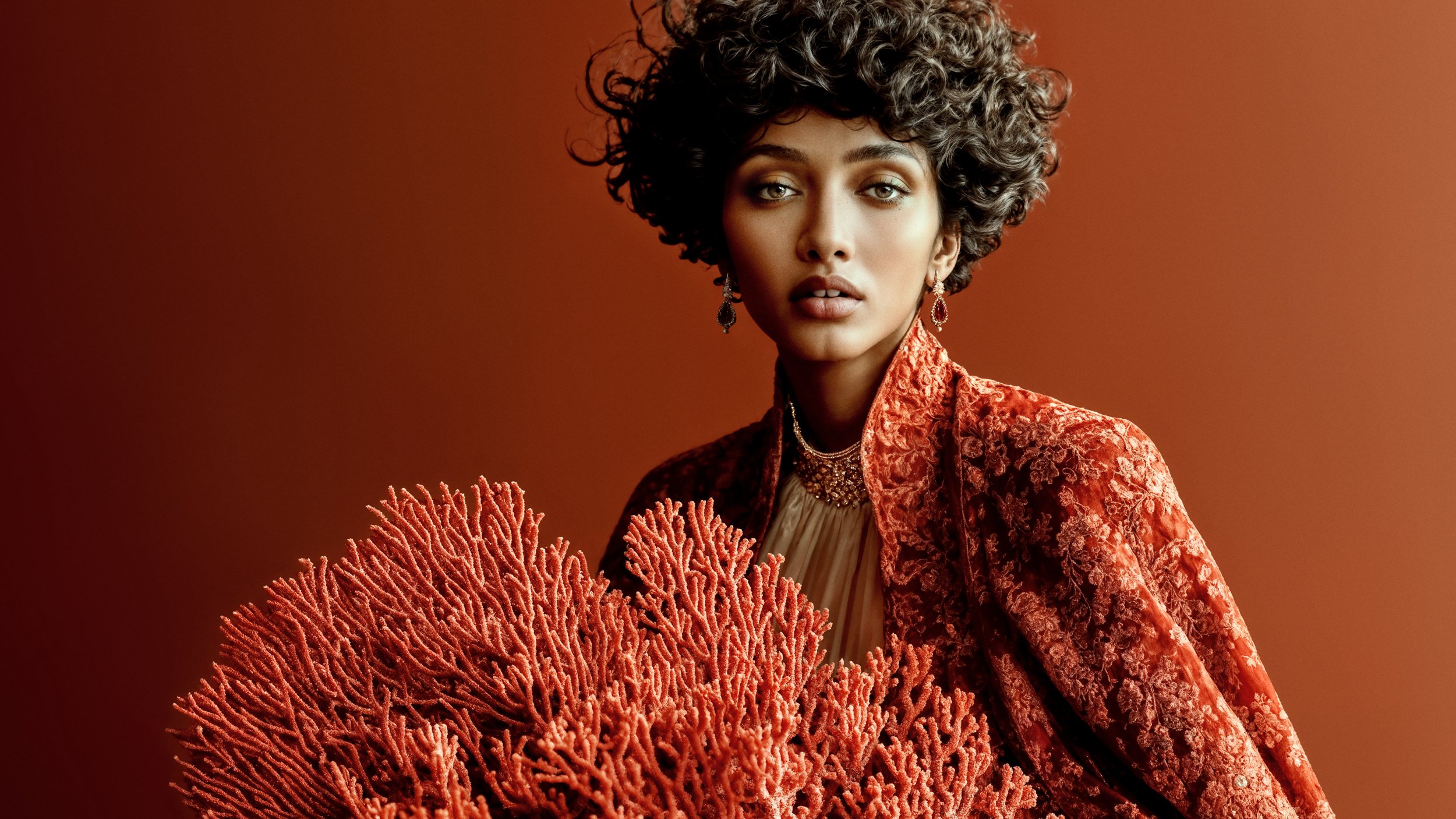 Kavya beside a large branching red coral formation, wearing richly textured red clothing against a warm terracotta background.