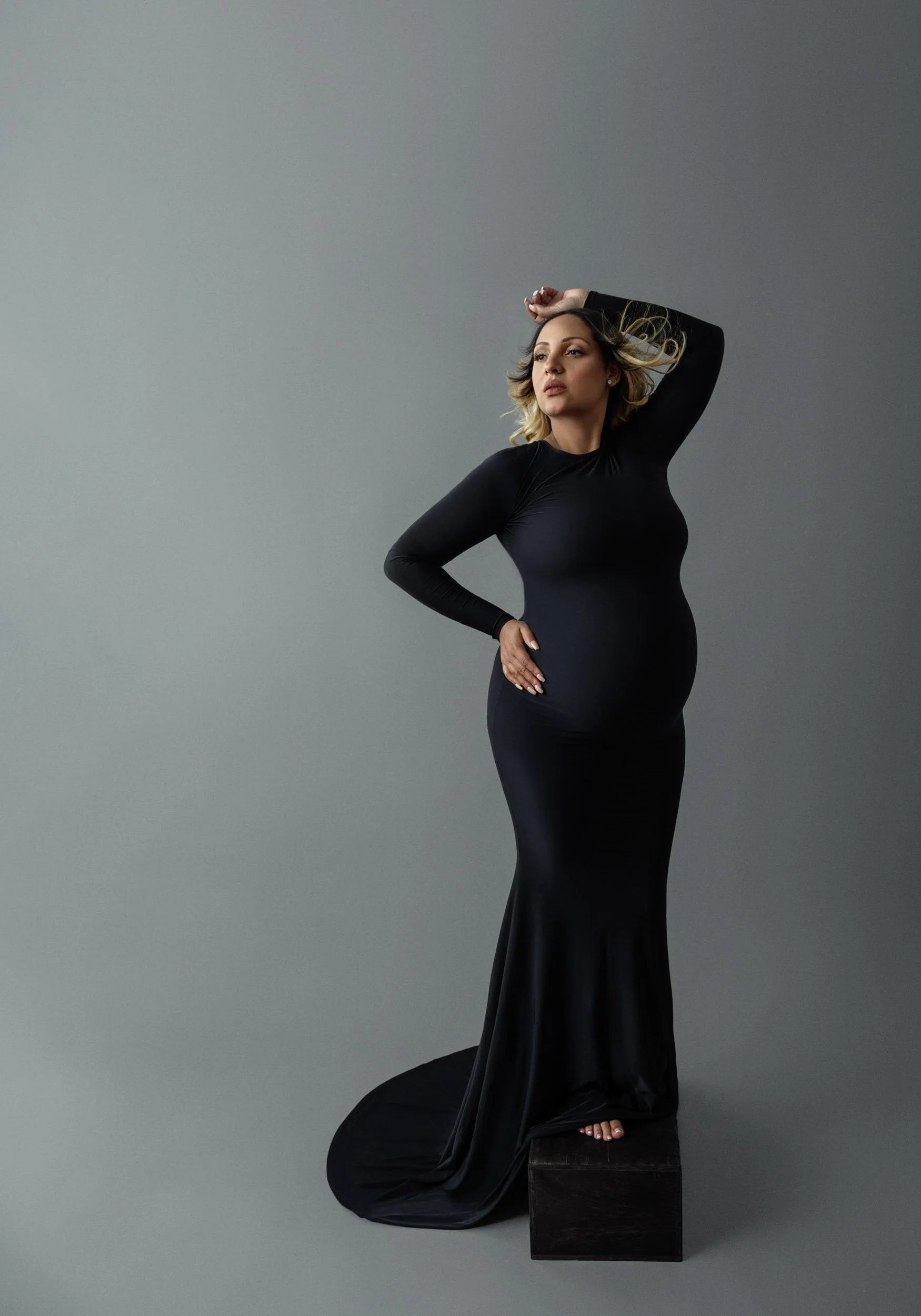 Full-length maternity portrait of a pregnant woman wearing a long black gown and wind blown hair against a gray studio background.