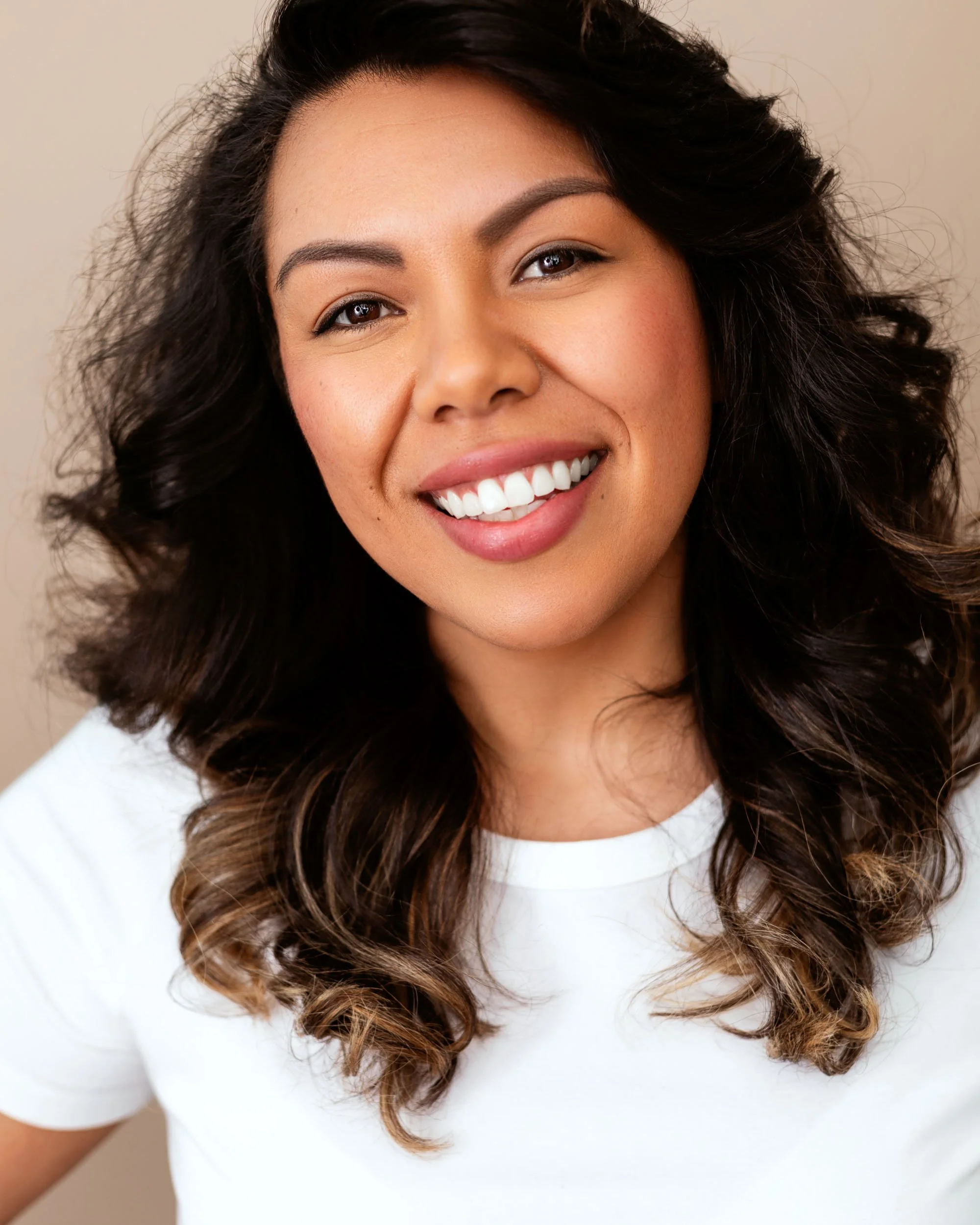 Personal branding headshot of a smiling woman with long wavy dark hair wearing a white top against a warm neutral background.