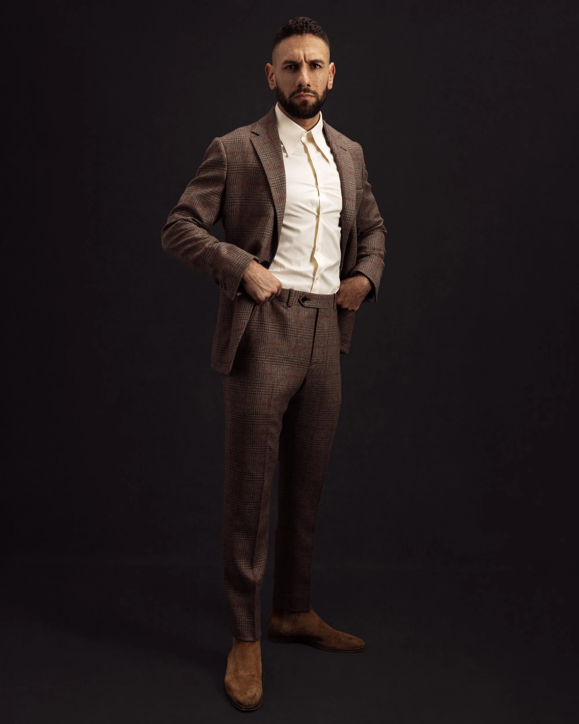 Full-length studio portrait of a man in a brown suit and white shirt standing against a dark neutral background.