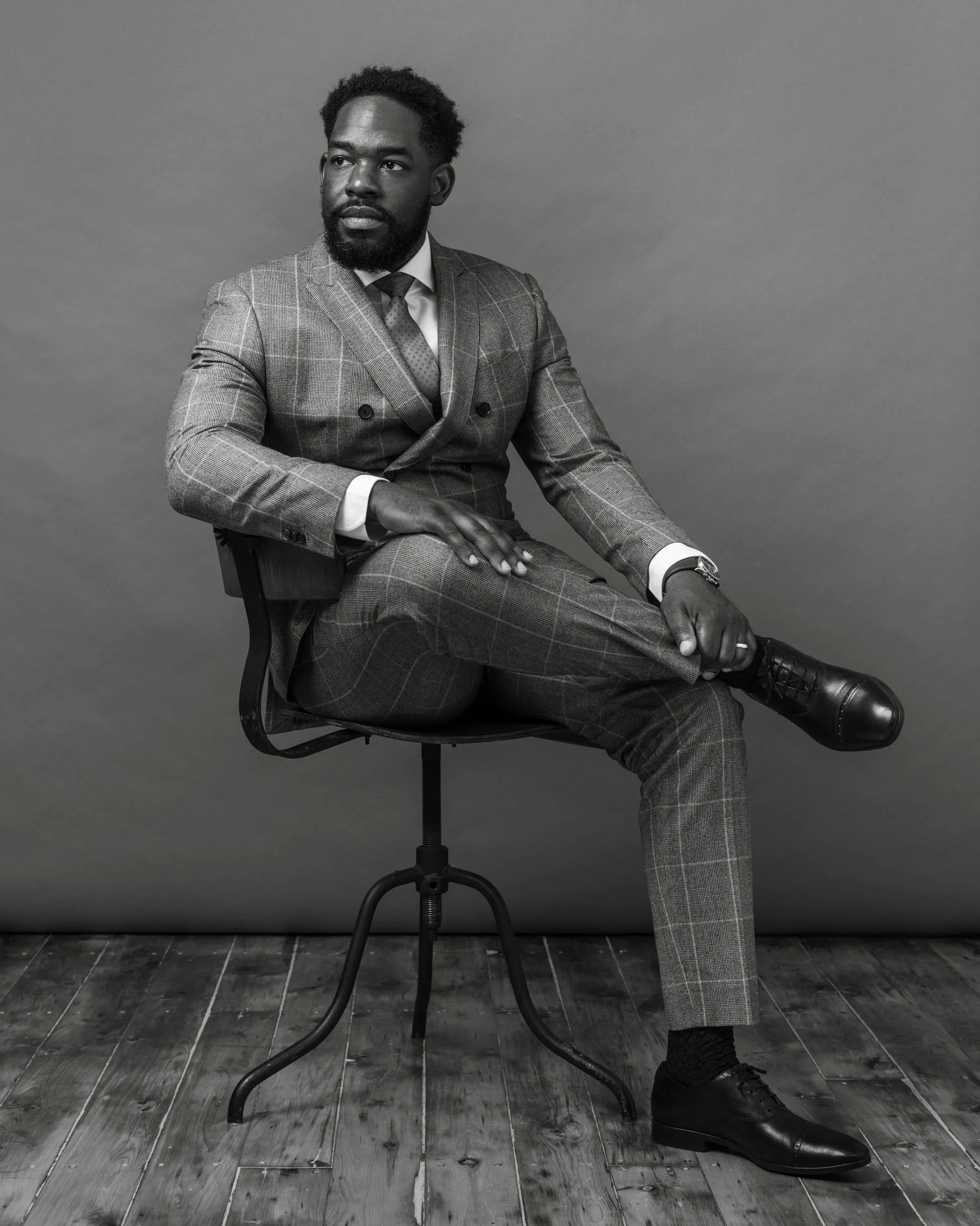 Black-and-white studio portrait of a seated man wearing a plaid suit and glasses against a plain gray backdrop.